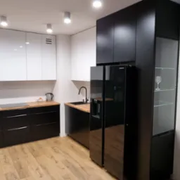 Nowoczesna kuchnia w czarno-białej kolorystyce z drewnianymi blatami, zabudowana szafka z przeszklonym frontem eksponująca kieliszek do wina, podłoga z paneli imitujących drewno.