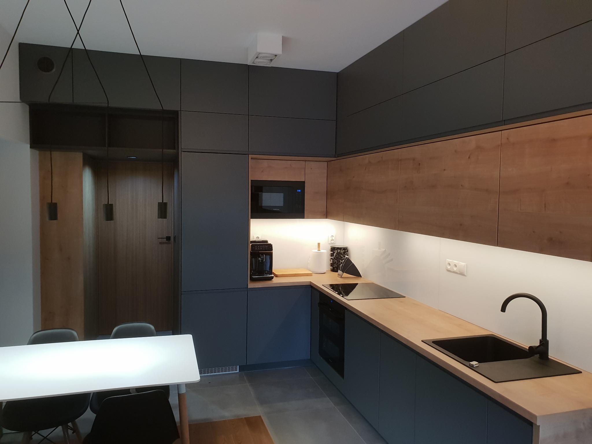 Nowoczesna kuchnia z szarymi i drewnianymi frontami, czarnym zlewem i płytą indukcyjną, oświetleniem podszafkowym i białym stołem z czarnymi krzesłami w tle.