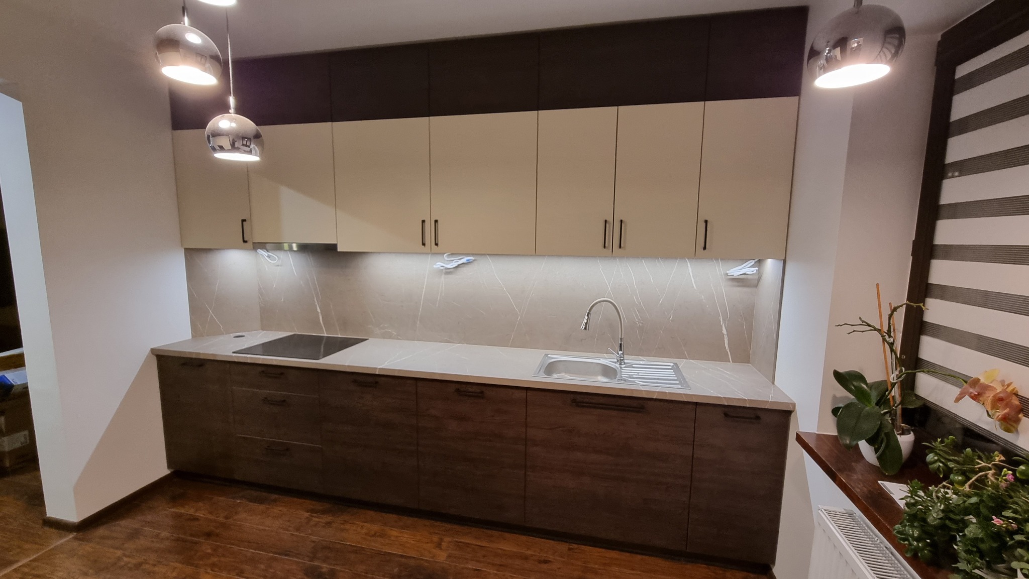 Nowoczesna kuchnia z dwukolorowymi szafkami (beżowe górne, brązowe dolne) i marmurkowym backsplash, oświetlona wiszącymi lampami i podświetleniem LED pod szafkami, z widocznymi elementami...