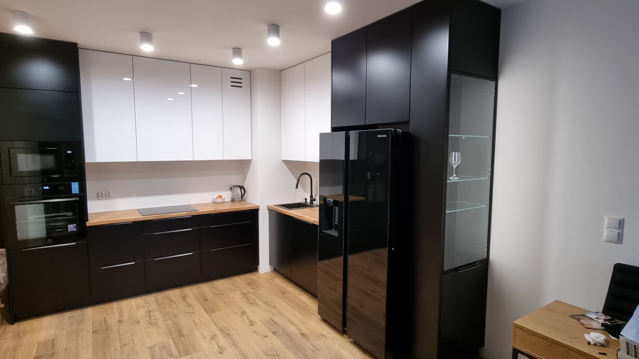 Nowoczesna kuchnia w czarno-białej kolorystyce z drewnianymi blatami, zabudowana szafka z przeszklonym frontem eksponująca kieliszek do wina, podłoga z paneli imitujących drewno.