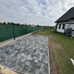 Ułożona szara kostka brukowa na posesji, obrzeża betonowe, zielona siatka ogrodzeniowa w tle, widoczny fragment domu.