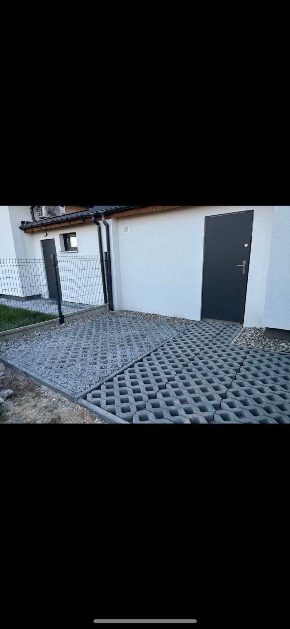 Szara ażurowa nawierzchnia ułożona z betonowych płyt przed budynkiem z szarymi drzwiami i metalowym ogrodzeniem.