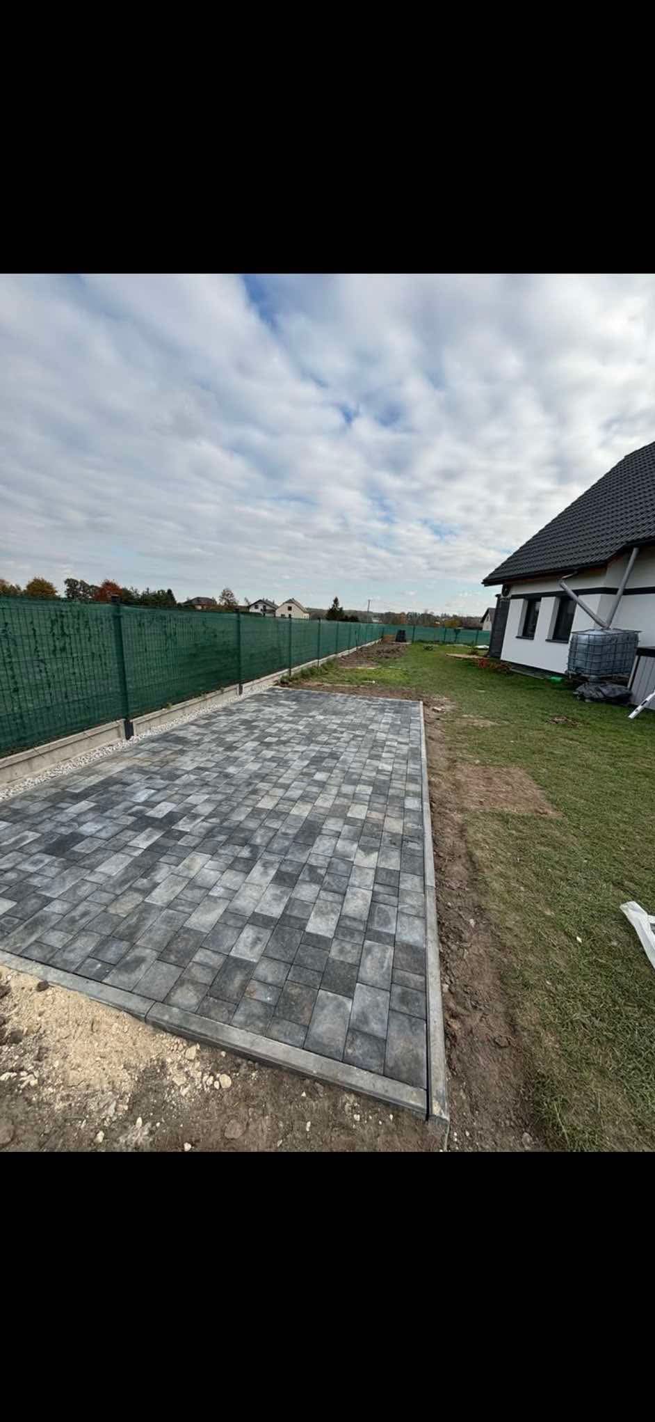 Ułożona szara kostka brukowa na posesji, obrzeża betonowe, zielona siatka ogrodzeniowa w tle, widoczny fragment domu.