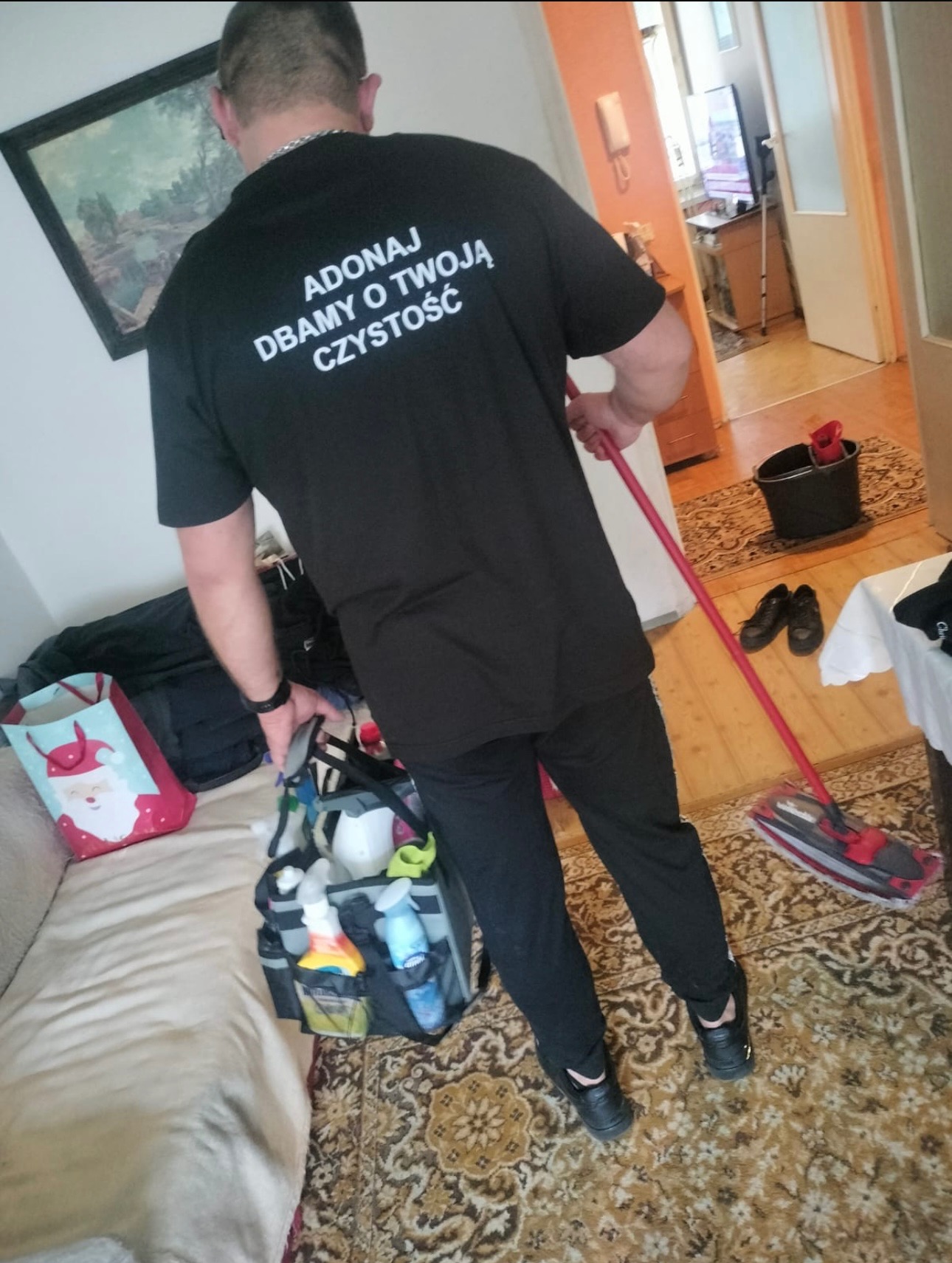 Osoba w czarnej koszulce z napisem sprzątająca podłogę mopem. Widoczna torba ze środkami czystości, wiadro i dywan w mieszkaniu. Perspektywa zza pleców.