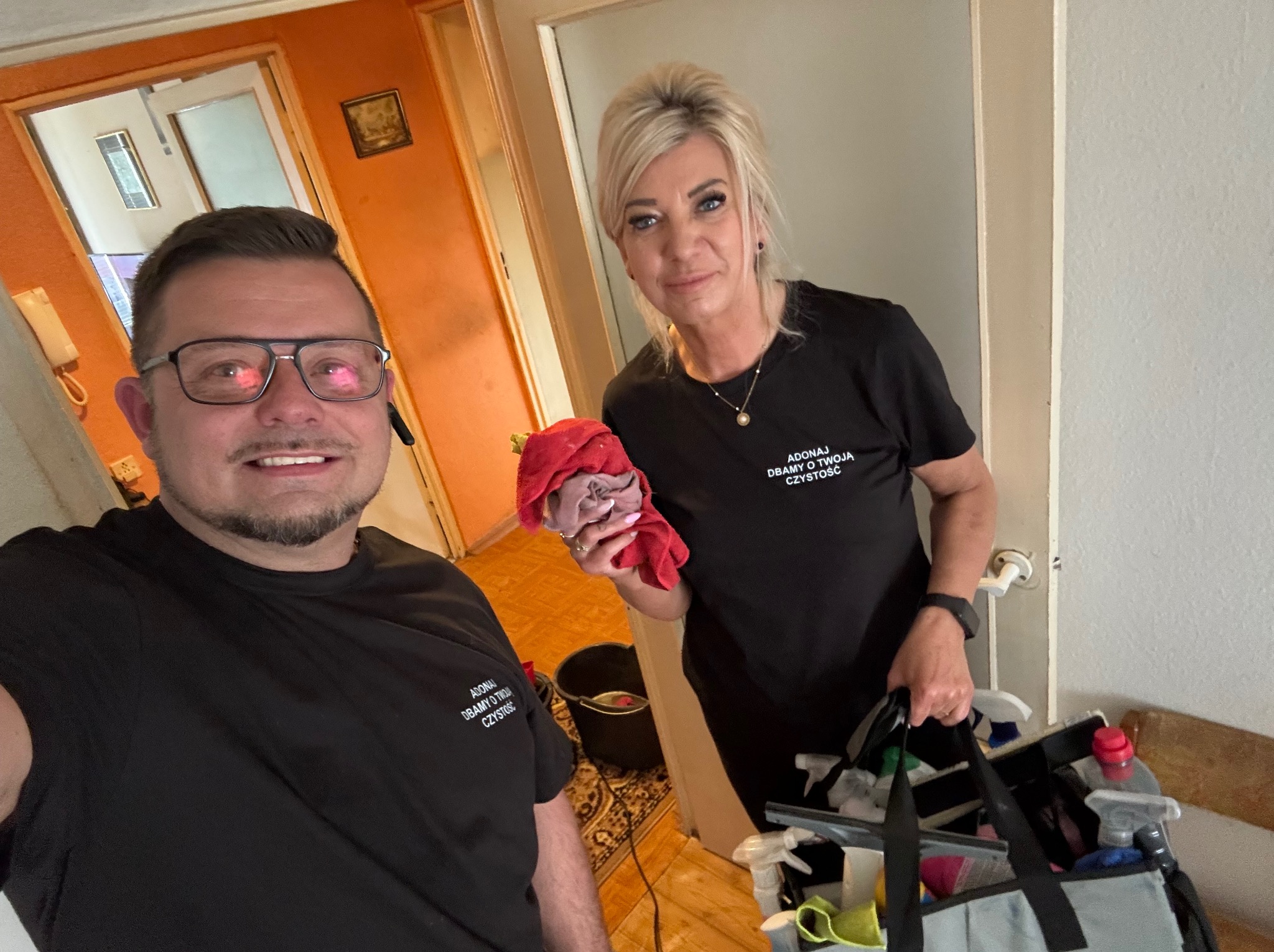 Uśmiechnięci pracownicy firmy sprzątającej w czarnych koszulkach z logo, trzymający wiadro i torbę ze środkami czystości, gotowi do pracy w pomarańczowym wnętrzu.