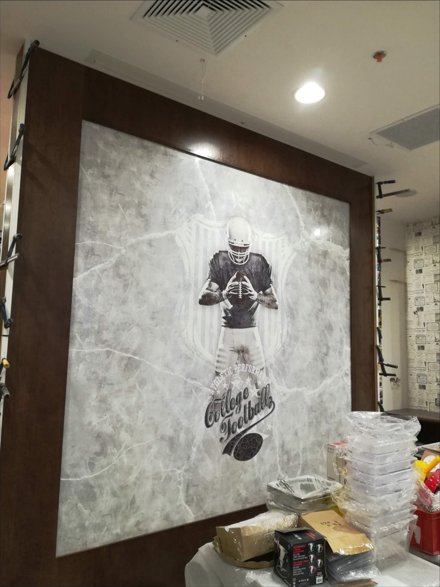 Ściana z dekoracyjnym wzorem imitującym marmur, oprawiona w ciemnobrązową ramę, przedstawiająca motyw futbolu amerykańskiego, widoczne akcesoria i materiały budowlane w pomieszczeniu.