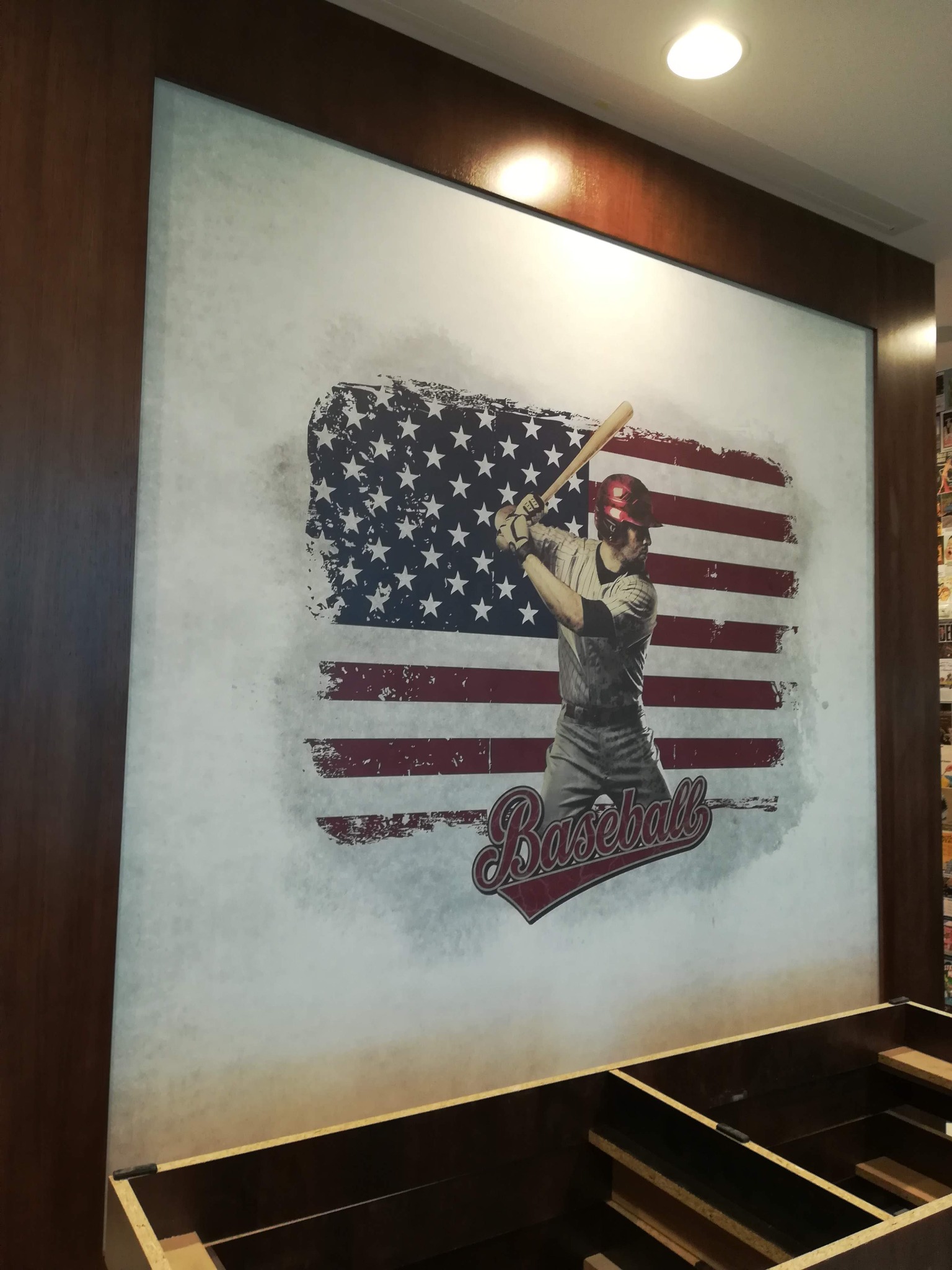 Duży fototapeta z motywem baseballisty na tle stylizowanej flagi USA, zamontowana na ścianie w lokalu, widoczne fragmenty drewnianej ramy i niedokończonej zabudowy pod ścianą.