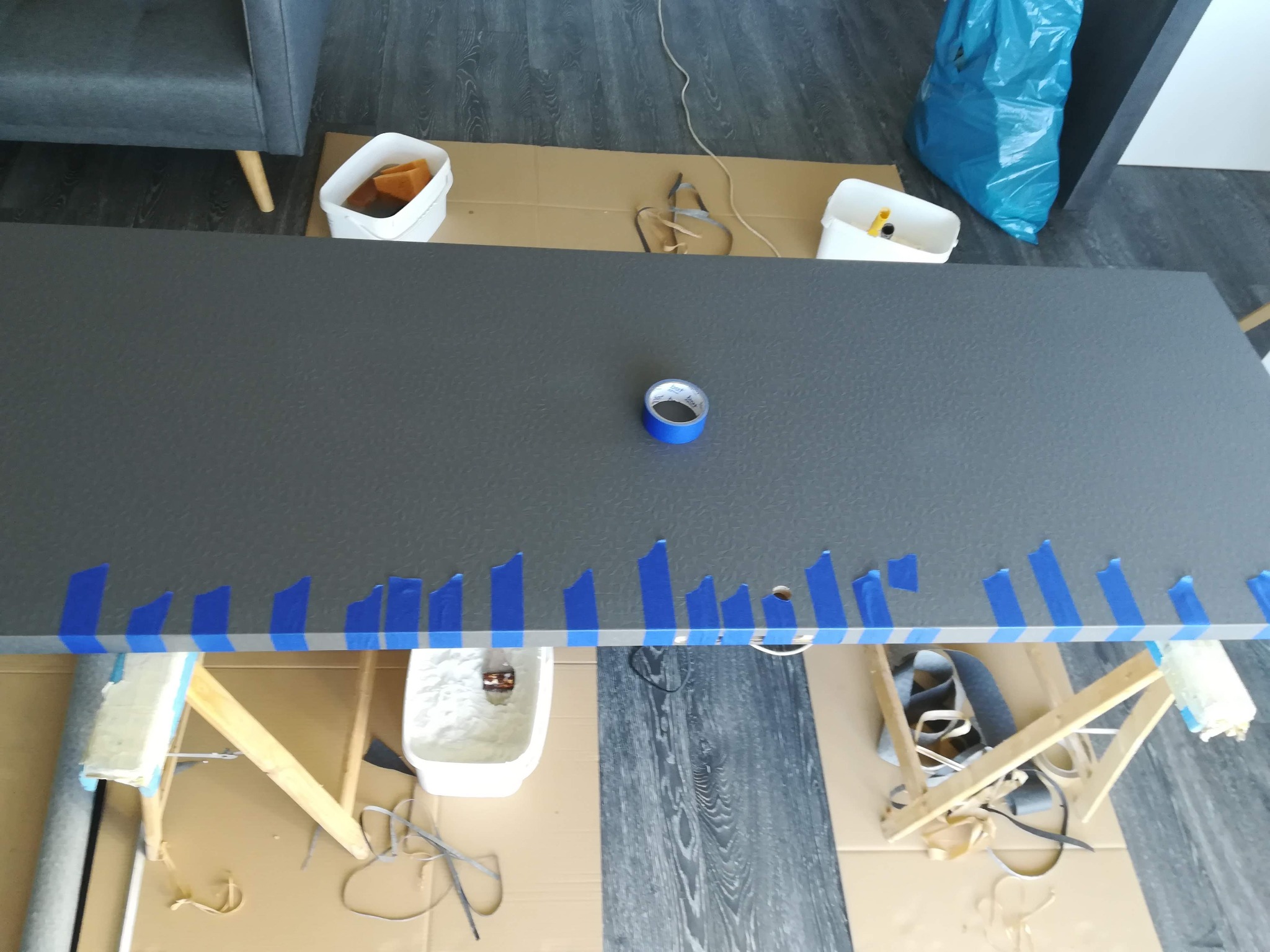 Przygotowanie do malowania: szary blat zabezpieczony niebieską taśmą malarską, widoczne wiadra z farbą, wałki i stojaki malarskie