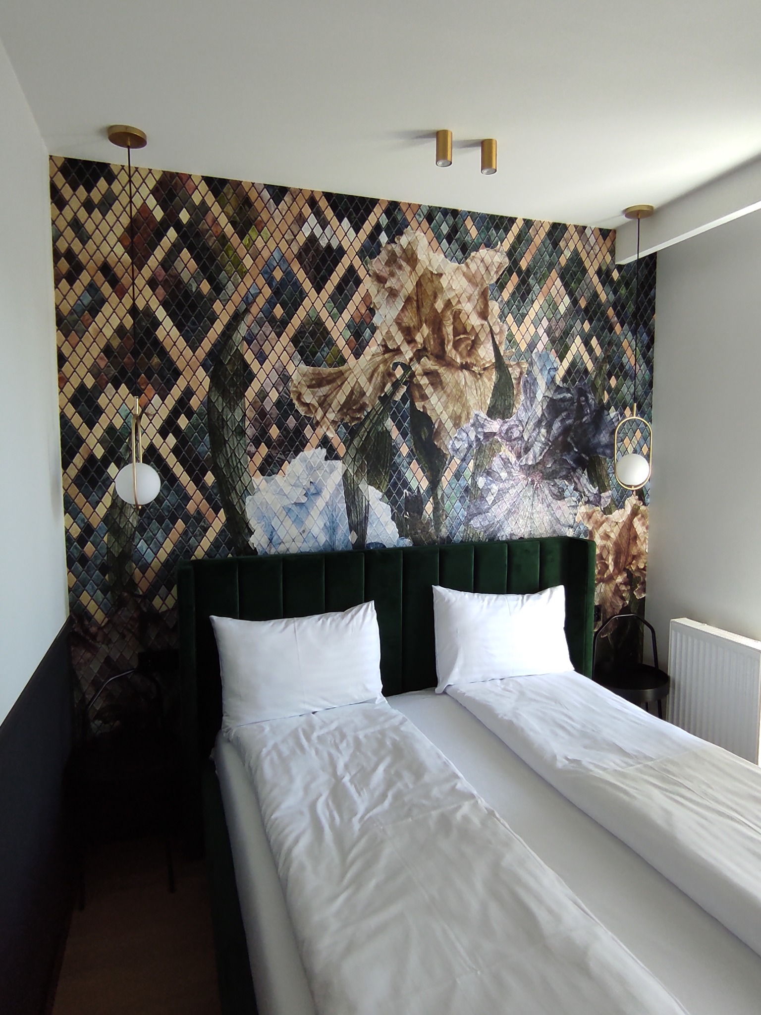 Sypialnia z łóżkiem dwuosobowym i dekoracyjną tapetą w romby z motywem kwiatowym, oświetlona złotymi lampami wiszącymi i sufitowymi.