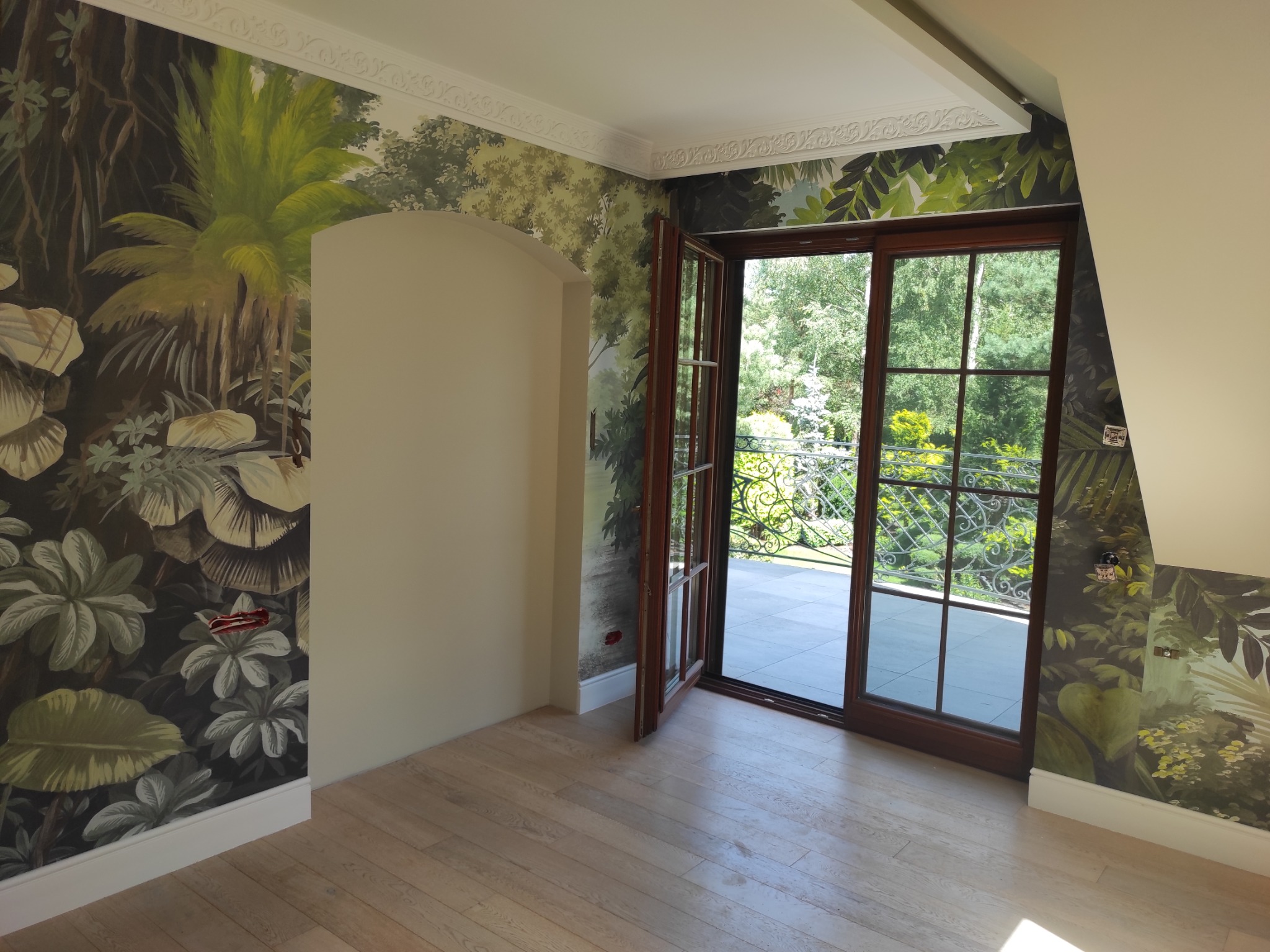 Pokój po odświeżeniu, ściany pokryte tapetą z motywem tropikalnego lasu, drewniana podłoga i otwarte drzwi balkonowe z widokiem na zieleń.