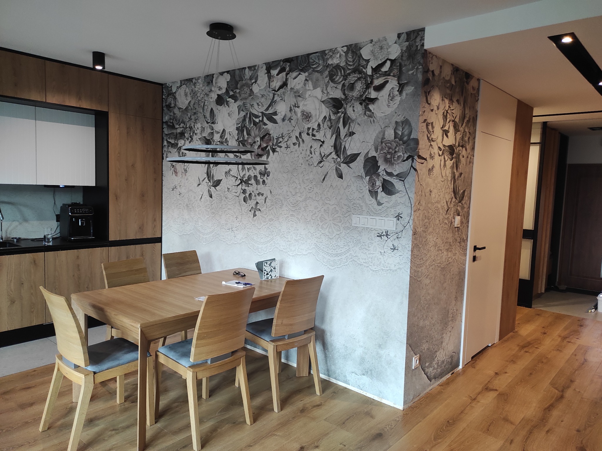 Jadalnia w Geffen po remoncie, ściana z fototapetą w kwiaty i koronki łączy się z fragmentem ściany w surowym stylu, drewniany stół i krzesła.