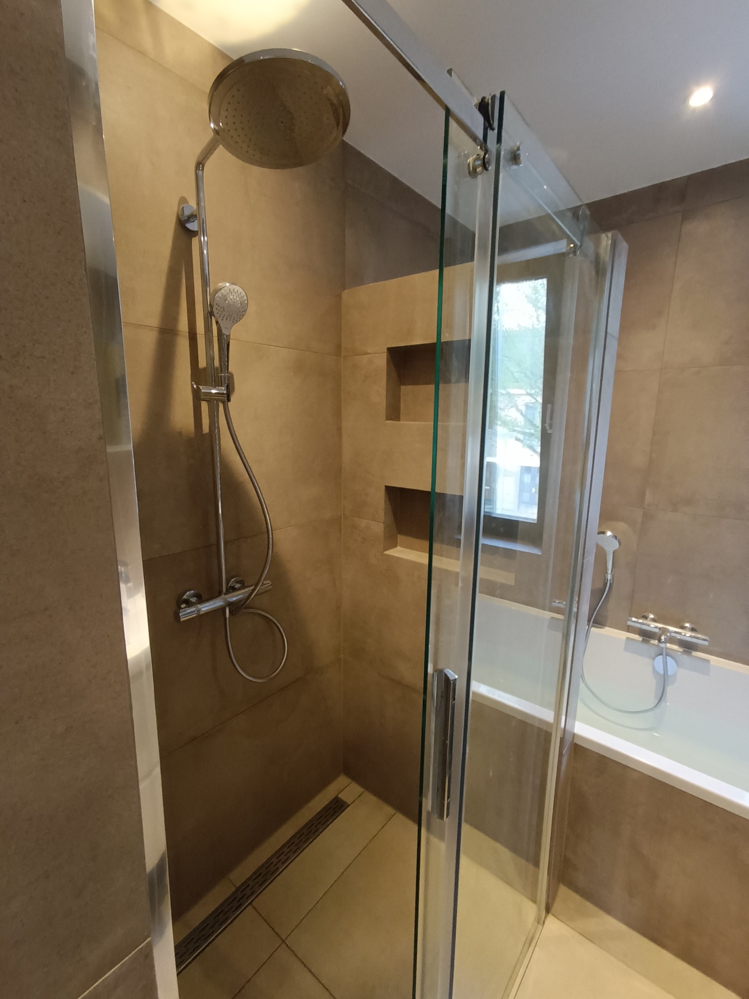 Nowoczesna kabina prysznicowa z deszczownicą i szklanymi drzwiami przesuwnymi, w łazience wykończonej płytkami w odcieniach beżu i brązu, z widocznym odpływem liniowym w podłodze.