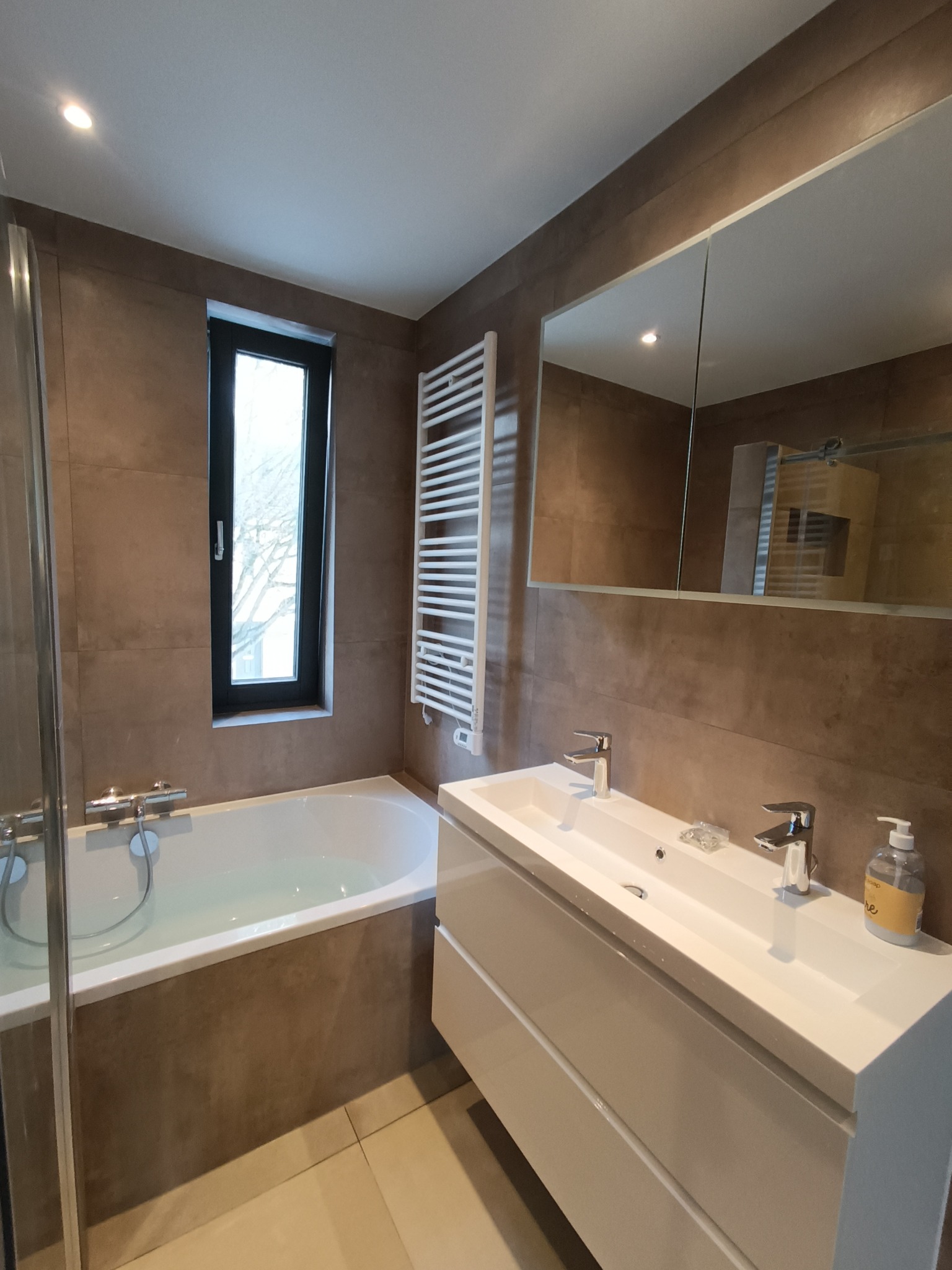 Nowoczesna łazienka z brązowymi płytkami, białą wanną wypełnioną wodą, grzejnikiem drabinkowym i podwójną umywalką z armaturą.