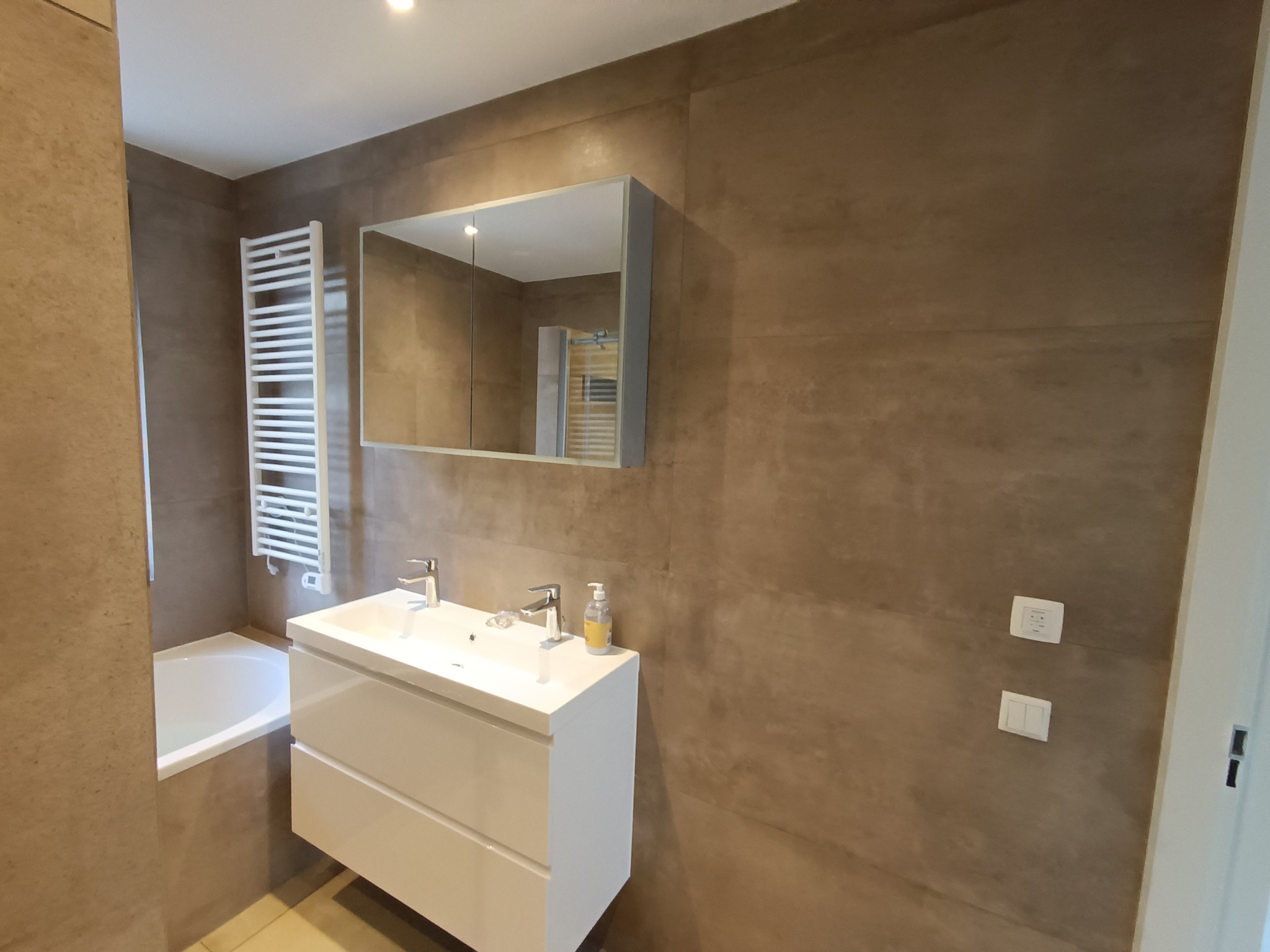 Nowoczesna łazienka z umywalką nablatową, lustrem w zabudowie i grzejnikiem drabinkowym na tle ścian wykończonych płytkami imitującymi beton.