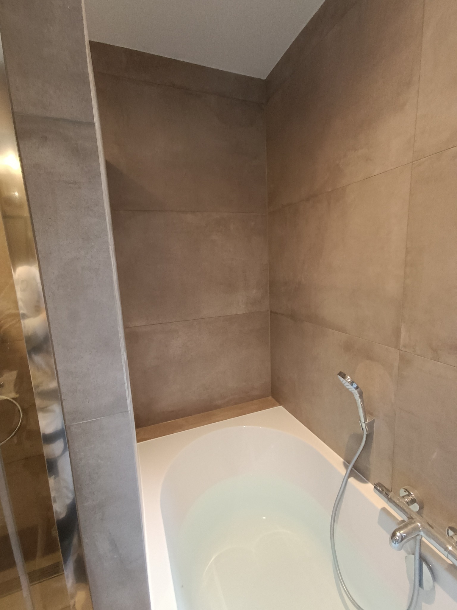 Wykończona łazienka z wanną, ściany wyłożone brązowymi płytkami imitującymi kamień, chromowana armatura i szklana kabina prysznicowa po lewej stronie.