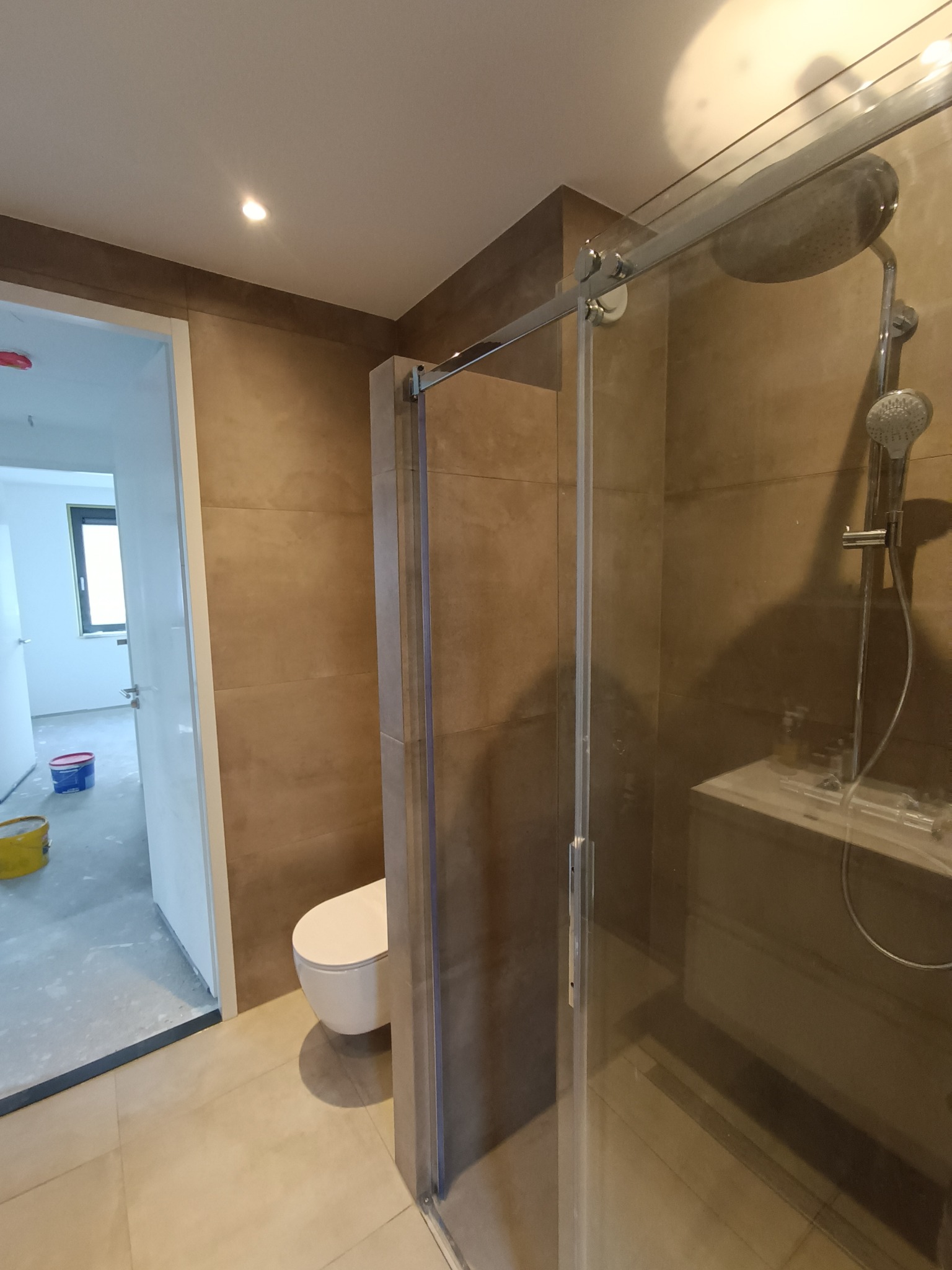 Nowoczesna kabina prysznicowa z deszczownicą w łazience wykończonej płytkami imitującymi kamień, widoczny fragment toalety i otwarte drzwi do pomieszczenia w stanie surowym z wiadrami farby.