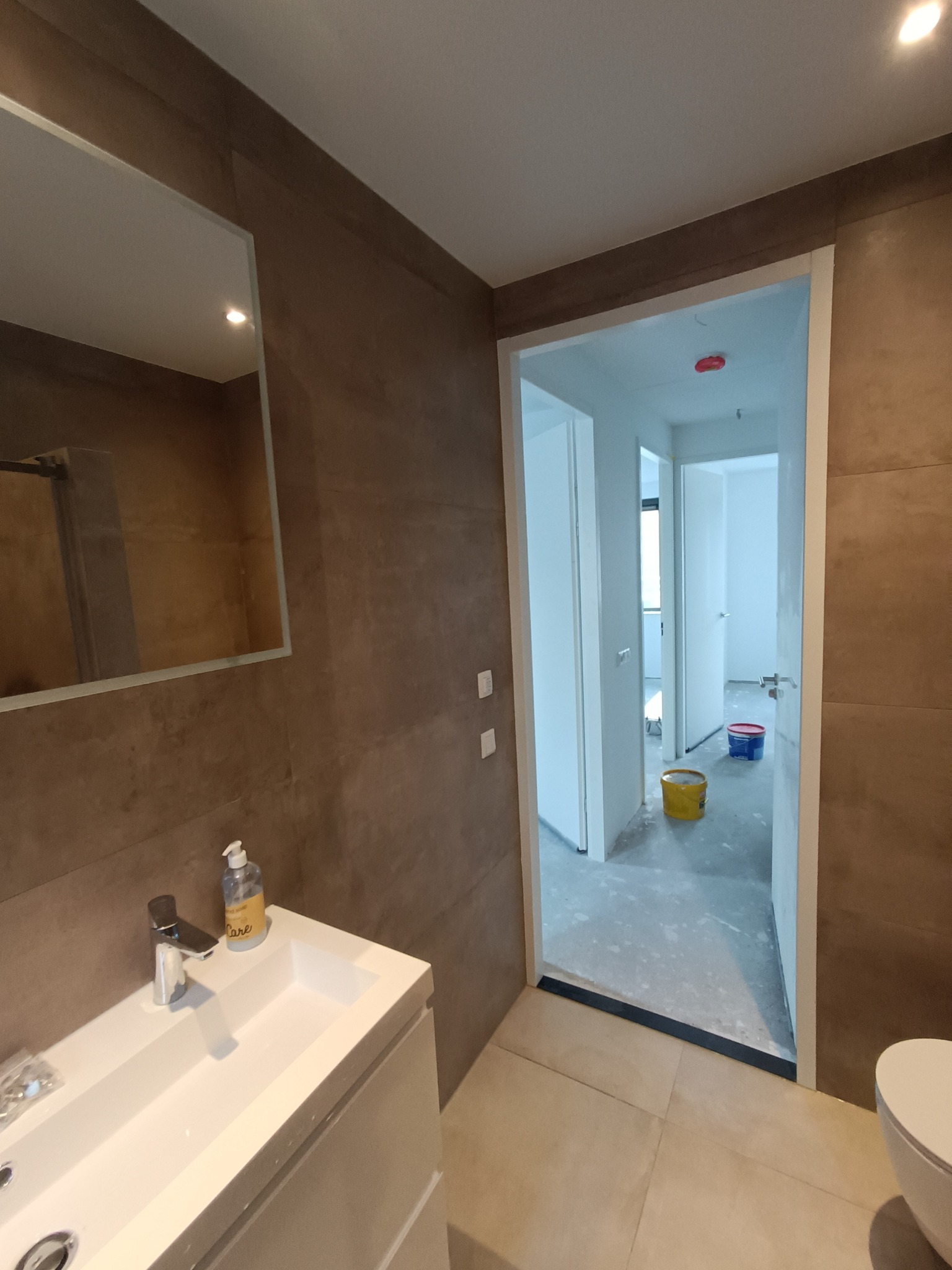 Nowoczesna łazienka w trakcie prac wykończeniowych: widoczna umywalka z baterią, duże płytki imitujące kamień na ścianach, lustro oraz otwarte drzwi do kolejnych pomieszczeń z wiadrami z farbą w tle.