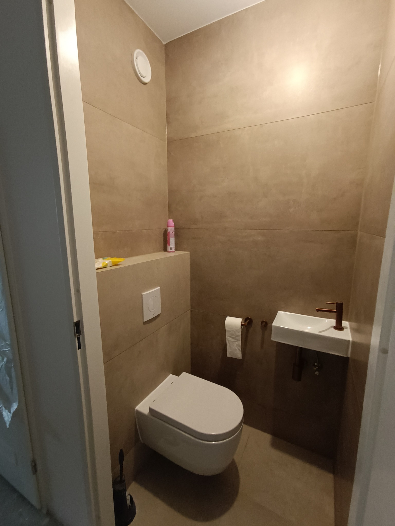 Nowoczesna toaleta z podwieszaną miską WC, umywalką nablatową z miedzianą armaturą i beżowymi płytkami imitującymi kamień na ścianach.