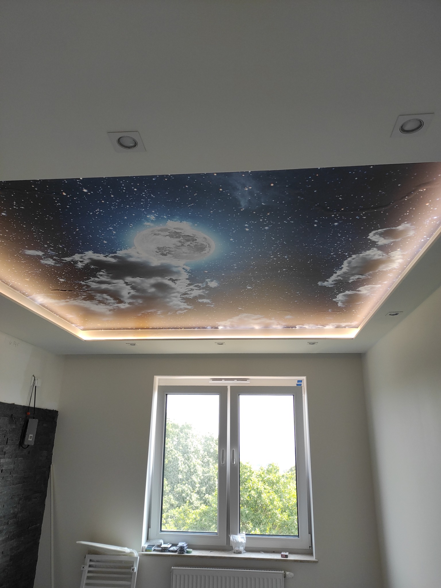 Sufit napinany z motywem nocnego nieba z księżycem i chmurami, podświetlony listwami LED, w pokoju z oknem i grzejnikiem.