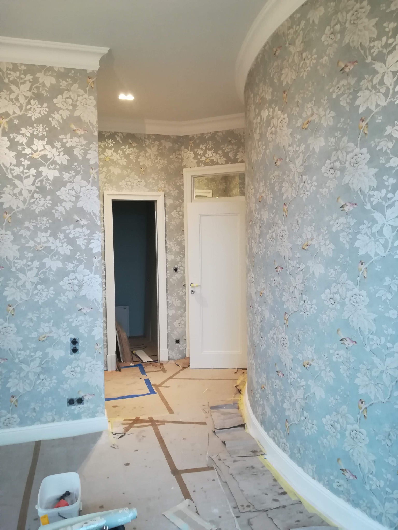 Wnętrze z niebieską tapetą w kwiaty i ptaki, widoczne narzędzia i przygotowania do prac wykończeniowych na podłodze, biała listwa przypodłogowa i sztukateria sufitowa.