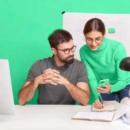 Dwoje grafików w okularach omawia projekt na smartfonie, notując szczegóły w notatniku na tle planów architektonicznych i monitora.