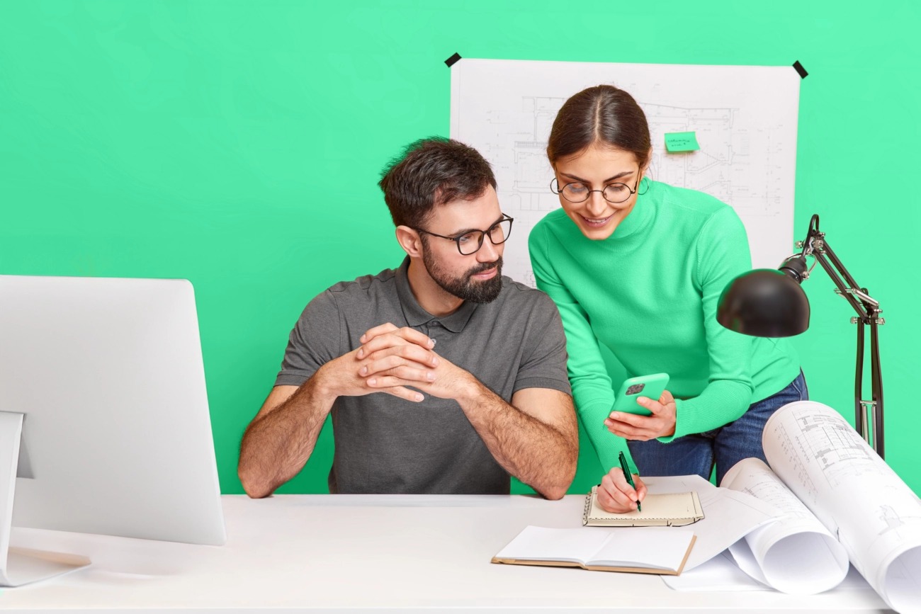 Dwoje grafików w okularach omawia projekt na smartfonie, notując szczegóły w notatniku na tle planów architektonicznych i monitora.