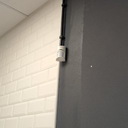 Federtech - Zamontowany czujnik ruchu na styku białej, kafelkowej ściany i szarej, gładkiej powierzchni. Czarny przewód instalacyjny biegnie wzdłuż ściany.