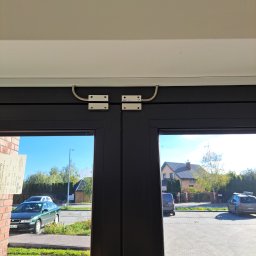 Federtech - Czujnik magnetyczny na czarnych drzwiach wejściowych. Widok na ulicę z domami i samochodami. Element systemu alarmowego zamontowany na futrynie.