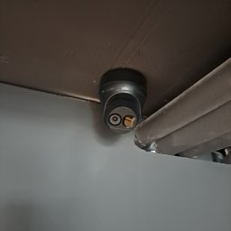 Federtech - Zamontowana czarna kamera monitoringu UAV pod sufitem, obok rur. Widok z dołu, bliska perspektywa. Szare ściany i ciemny sufit.