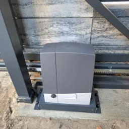 Szary automat do bram przesuwnych marki CAME, zamontowany na betonowej podstawie przy metalowej bramie z zębatą listwą, widoczny z bliska w dziennym świetle.