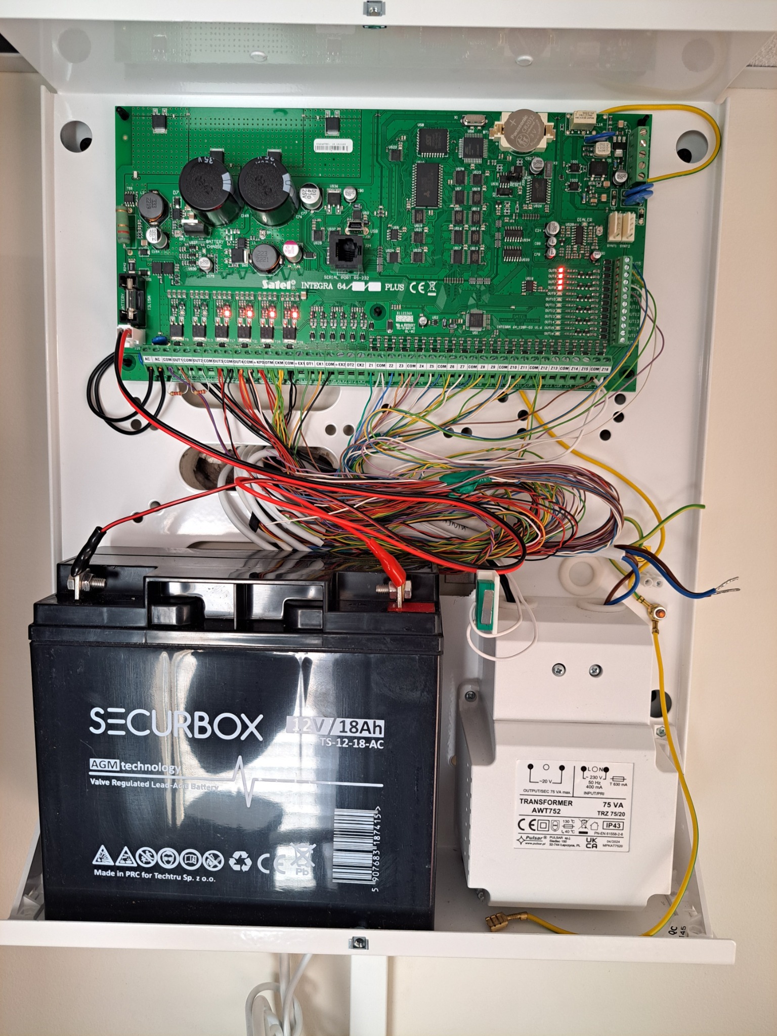 Panel alarmowy Satel Integra 64 Plus z podłączonym akumulatorem Securbox 12V/18Ah i transformatorem Pulsar AWT752, widoczne kable i elektronika.