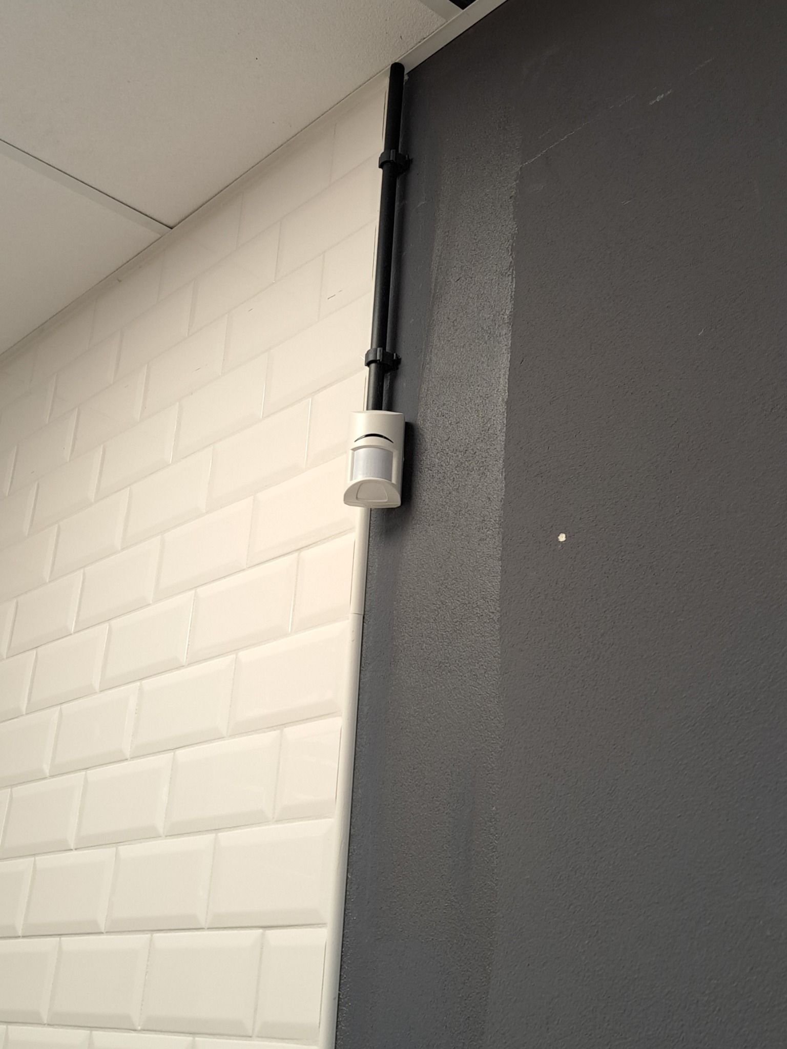 Zamontowany czujnik ruchu na styku białej, kafelkowej ściany i szarej, gładkiej powierzchni. Czarny przewód instalacyjny biegnie wzdłuż ściany.