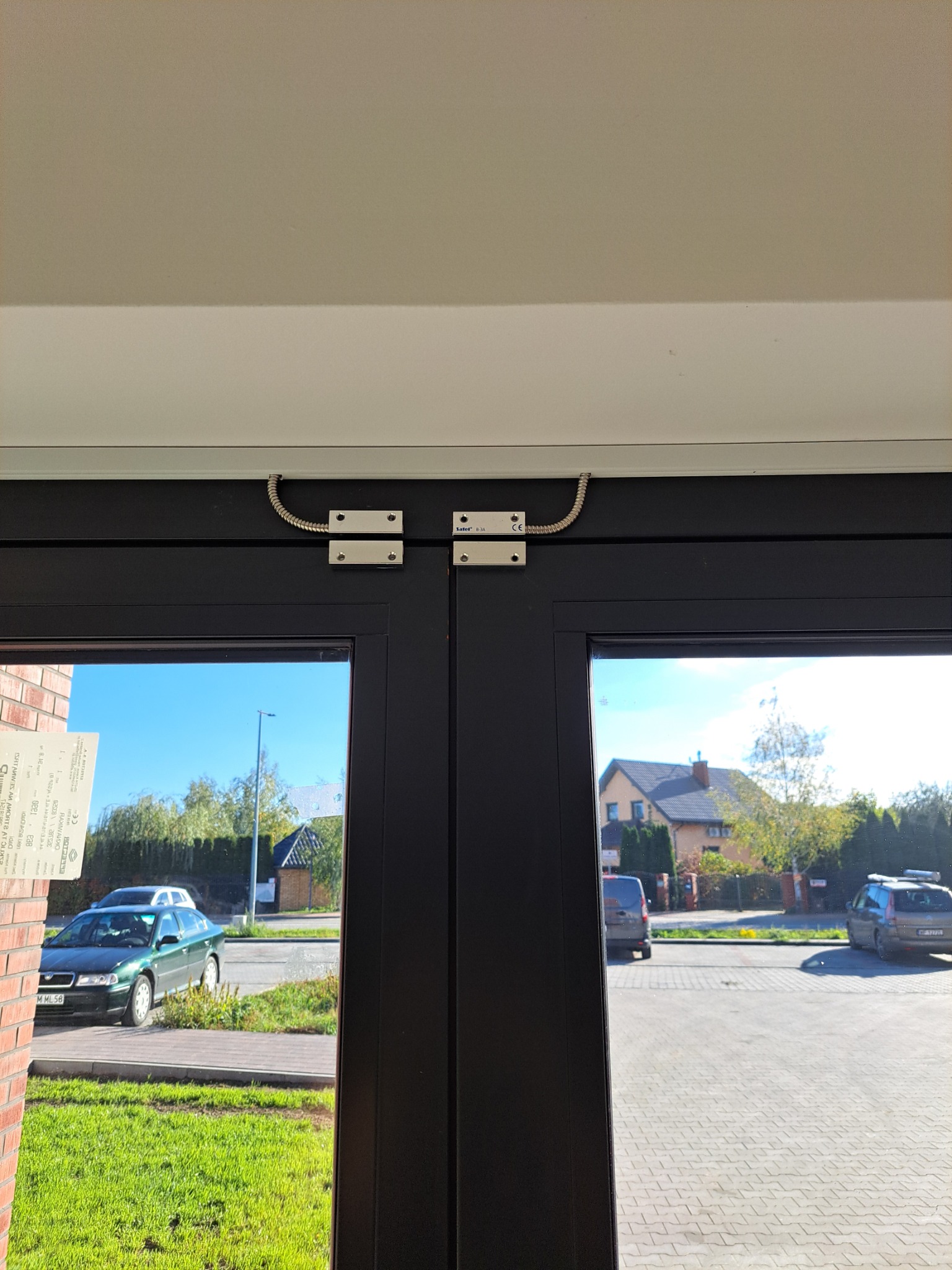 Czujnik magnetyczny na czarnych drzwiach wejściowych. Widok na ulicę z domami i samochodami. Element systemu alarmowego zamontowany na futrynie.