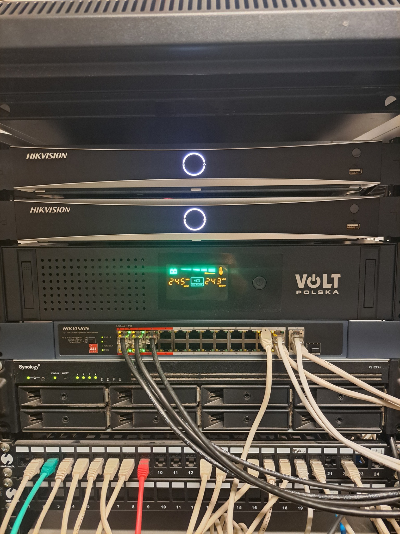 Szafa rack z urządzeniami Hikvision, Volt Polska i Synology. Widoczne kable sieciowe, switche i rejestratory. Elementy systemu monitoringu i zasilania awaryjnego.