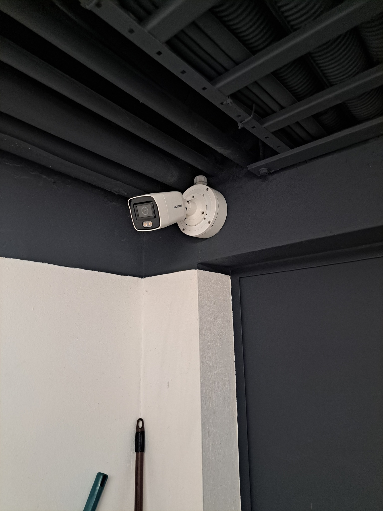 Biała kamera Hikvision zamontowana na ciemnej ścianie pod sufitem z widocznymi rurami. Obok framuga drzwi i fragment ściany. Ujęcie z dołu.