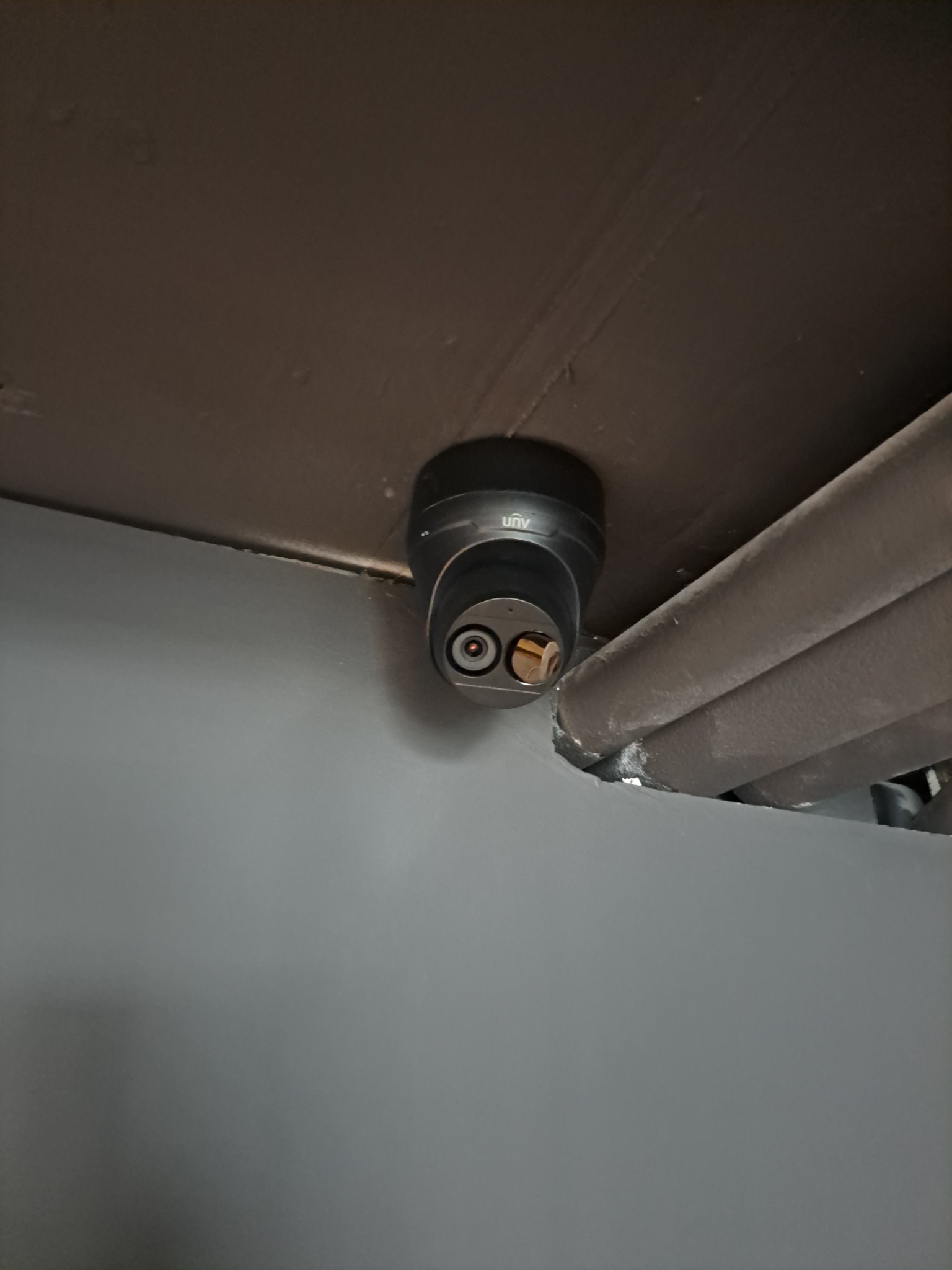 Zamontowana czarna kamera monitoringu UAV pod sufitem, obok rur. Widok z dołu, bliska perspektywa. Szare ściany i ciemny sufit.