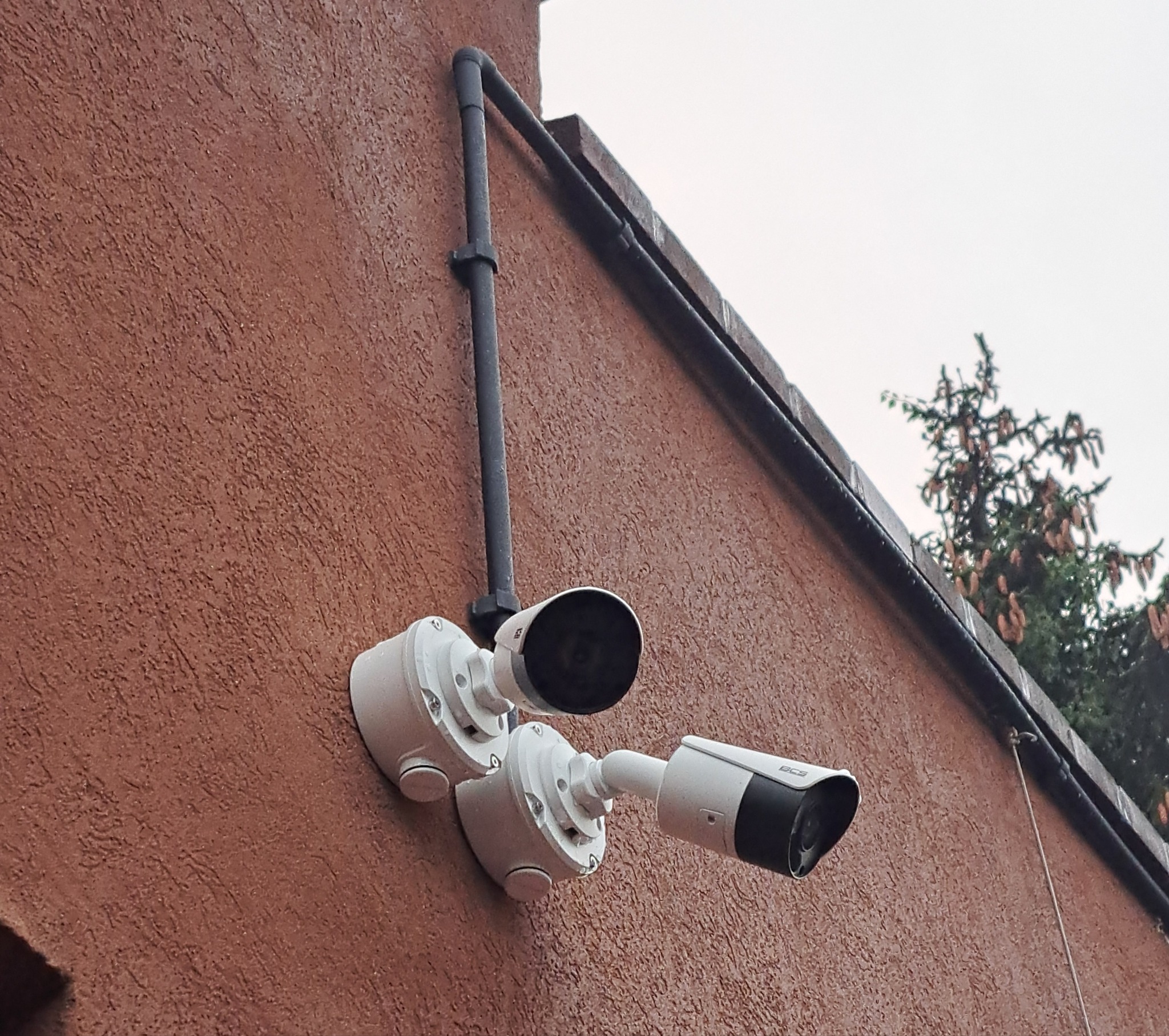 Dwie kamery monitoringu BCS zamontowane na brązowej, chropowatej ścianie budynku. Widoczne przewody i fragment iglastego drzewa w tle. Kątowe ujęcie.