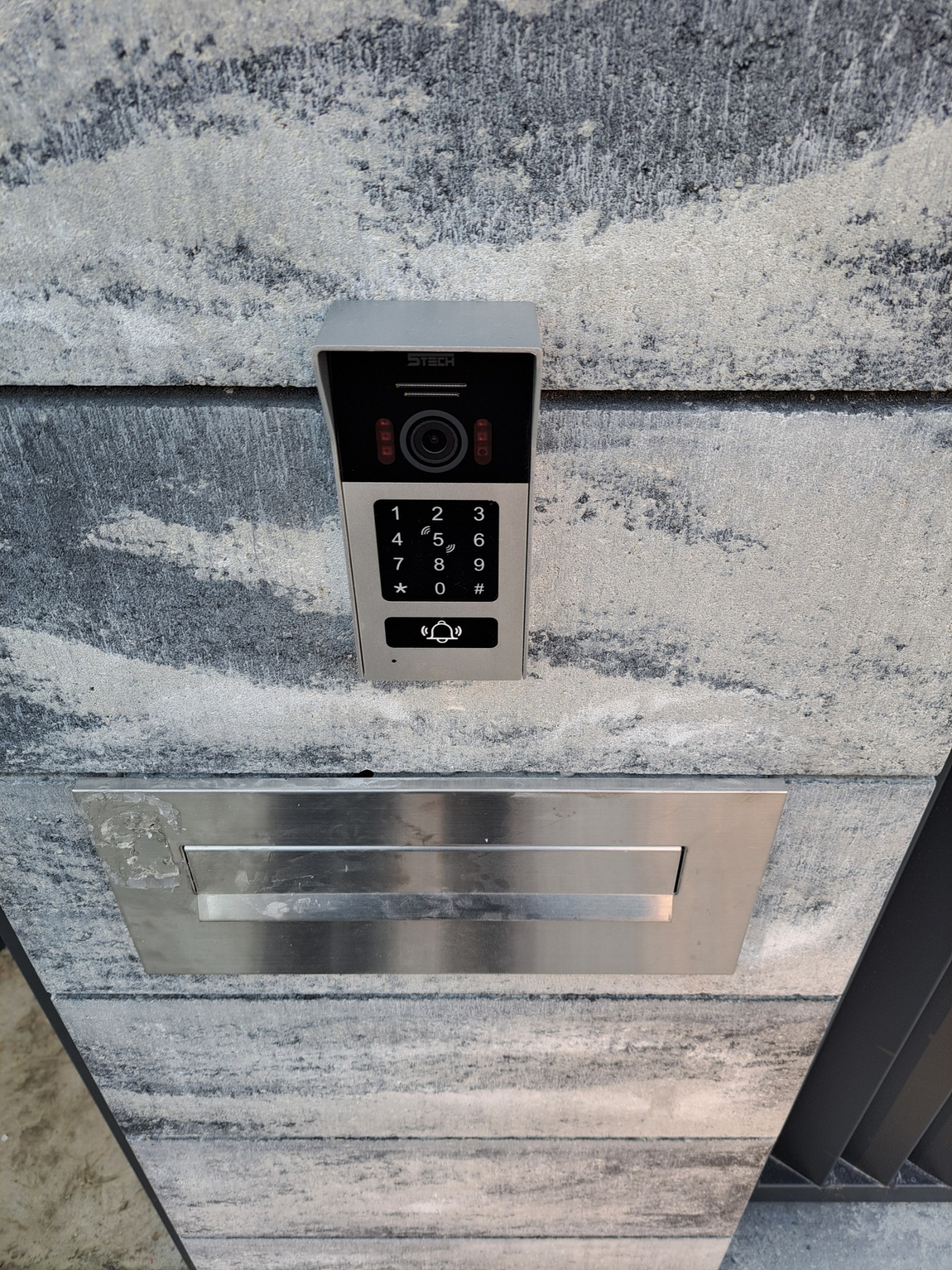 Nowoczesny wideodomofon z klawiaturą numeryczną i kamerą, zamontowany na szarej, teksturowanej elewacji obok skrzynki na listy.