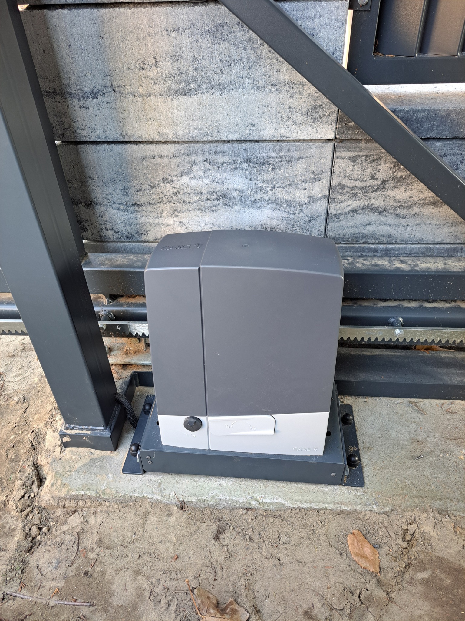 Szary automat do bram przesuwnych marki CAME, zamontowany na betonowej podstawie przy metalowej bramie z zębatą listwą, widoczny z bliska w dziennym świetle.