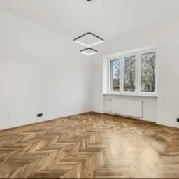 Puste, jasne wnętrze z parkietem w jodełkę, nowoczesnym oświetleniem w kształcie kwadratów i widokiem na budynek przez okno.