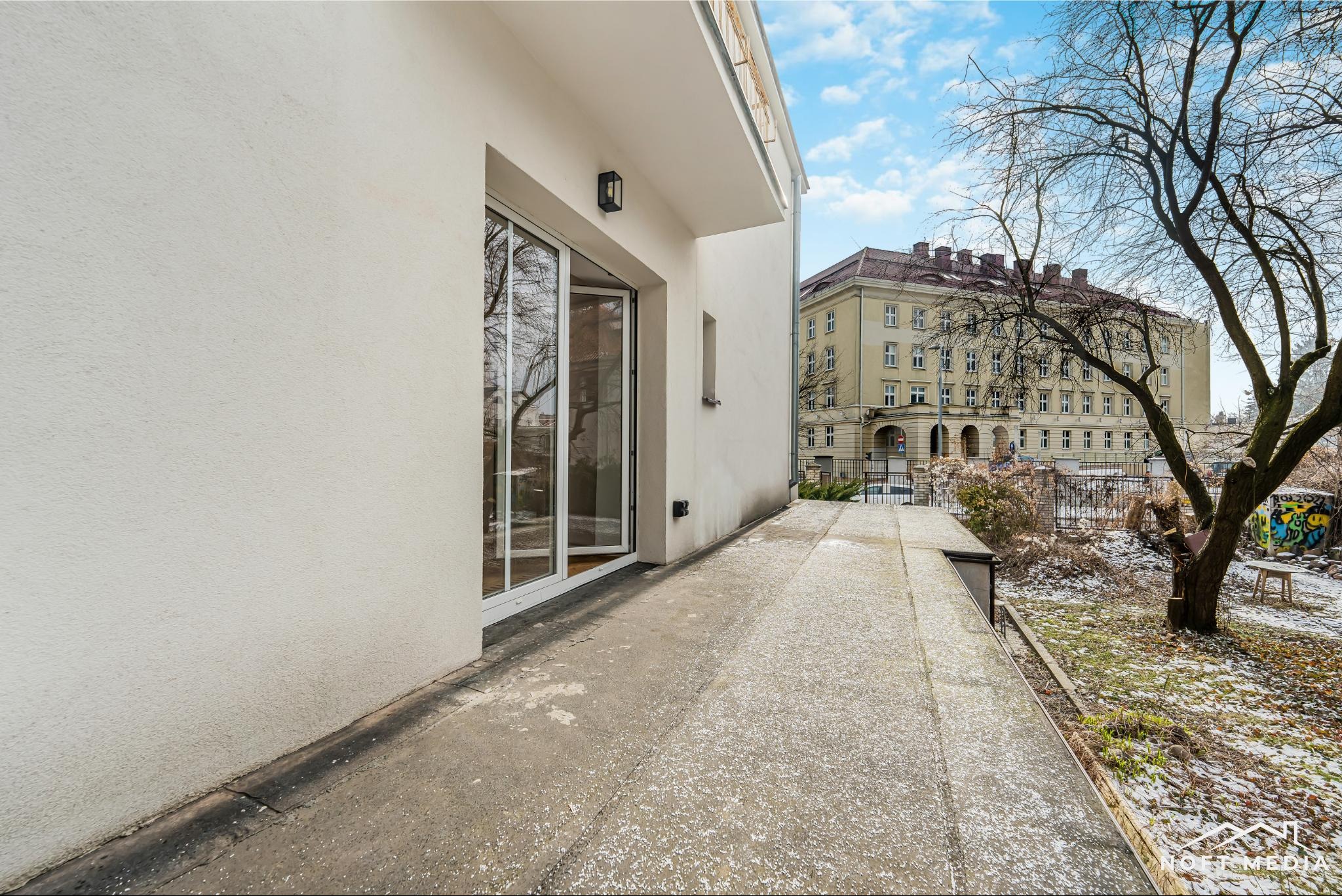 Nowoczesny balkon z otwartymi drzwiami balkonowymi, widok na budynek mieszkalny i ogród w zimowej scenerii.