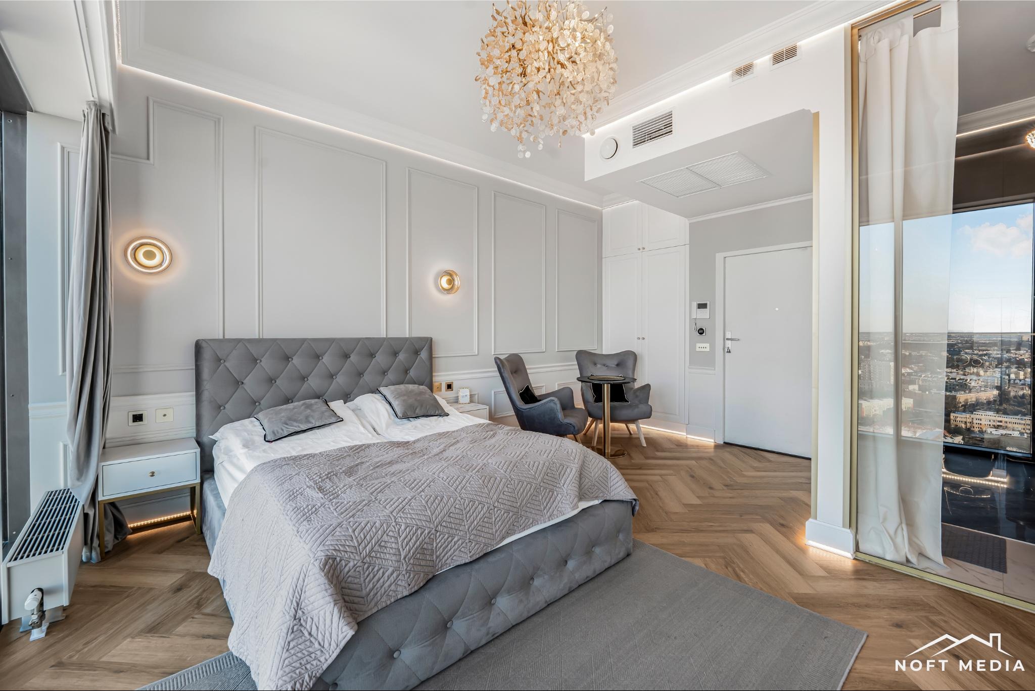 Elegancka sypialnia w odcieniach szarości z parkietem w jodełkę, ozdobnym żyrandolem i widokiem na miasto z wysokiego okna.