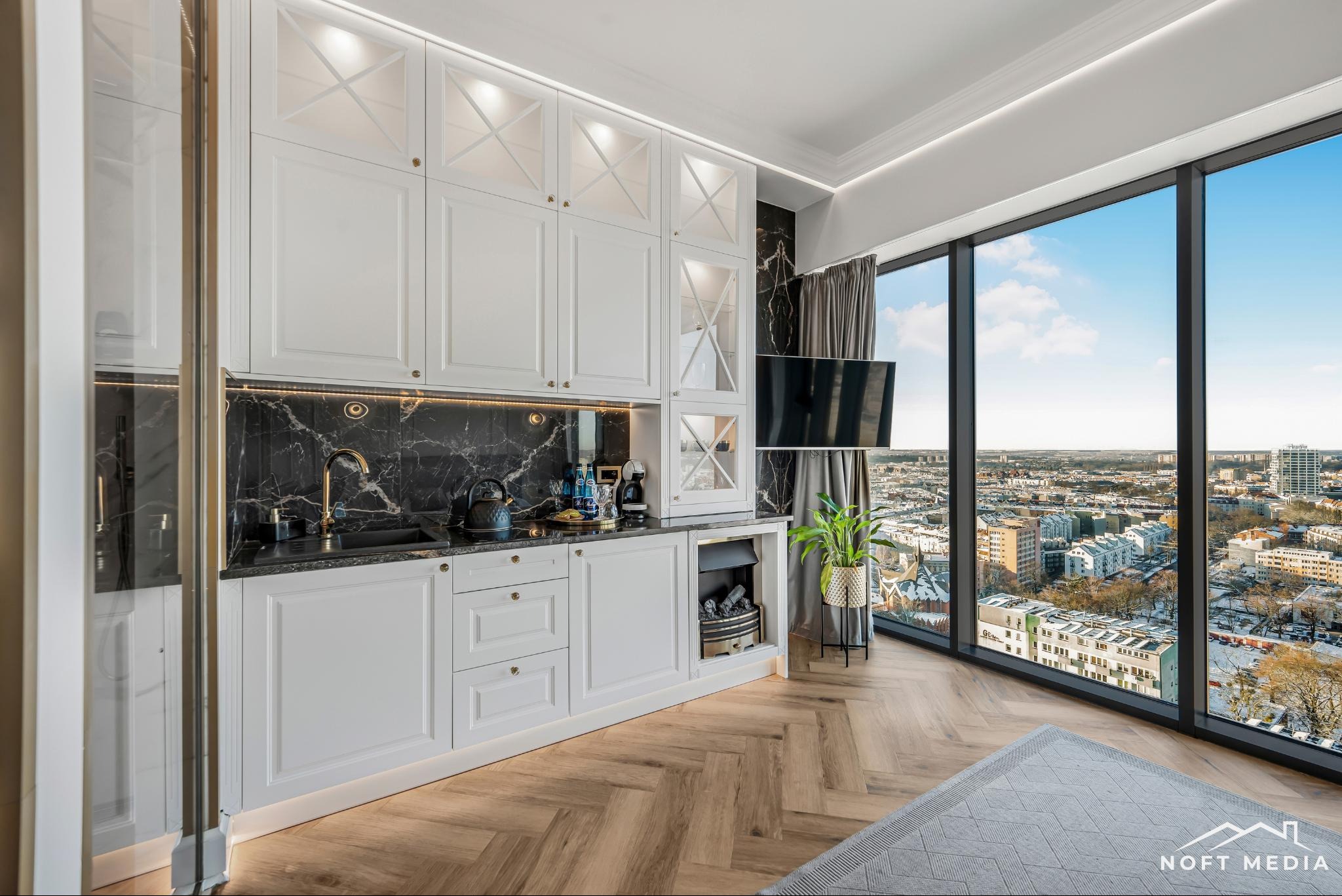 Nowoczesna kuchnia z białymi szafkami z przeszkleniami, czarnym marmurowym blatem i backsplashiem, złotą armaturą, widokiem na miasto przez panoramiczne okno i parkietem w jodełkę.