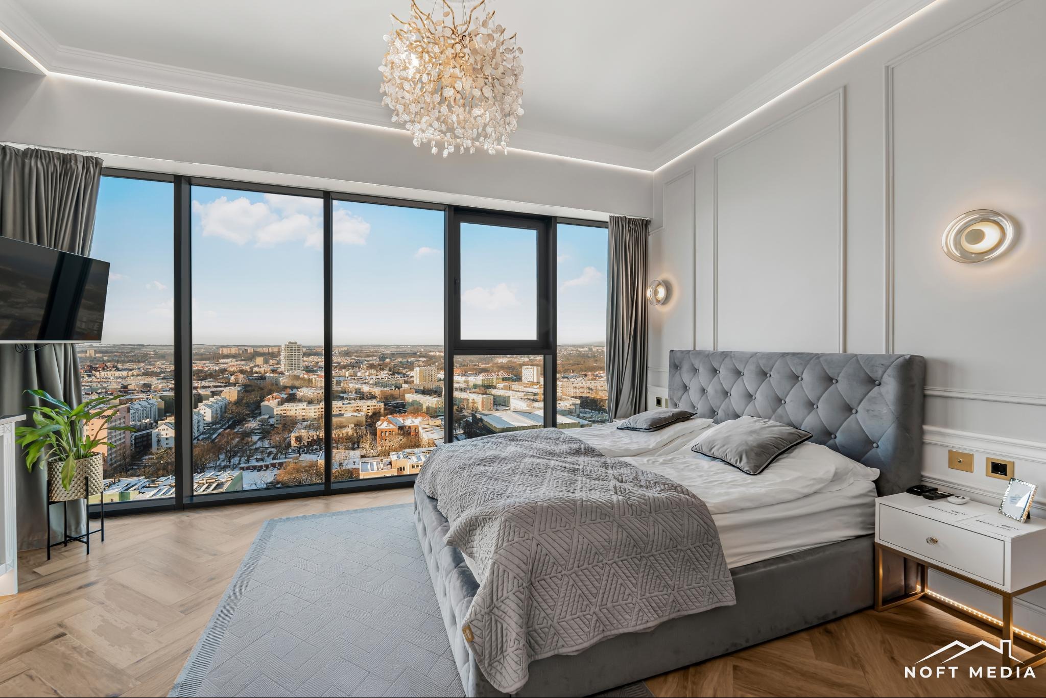 Elegancka sypialnia z dużym oknem panoramicznym ukazującym widok na miasto, szare łóżko z pikowanym zagłówkiem, kryształowy żyrandol, parkiet w jodełkę i białe ściany z dekoracyjnymi listwami.