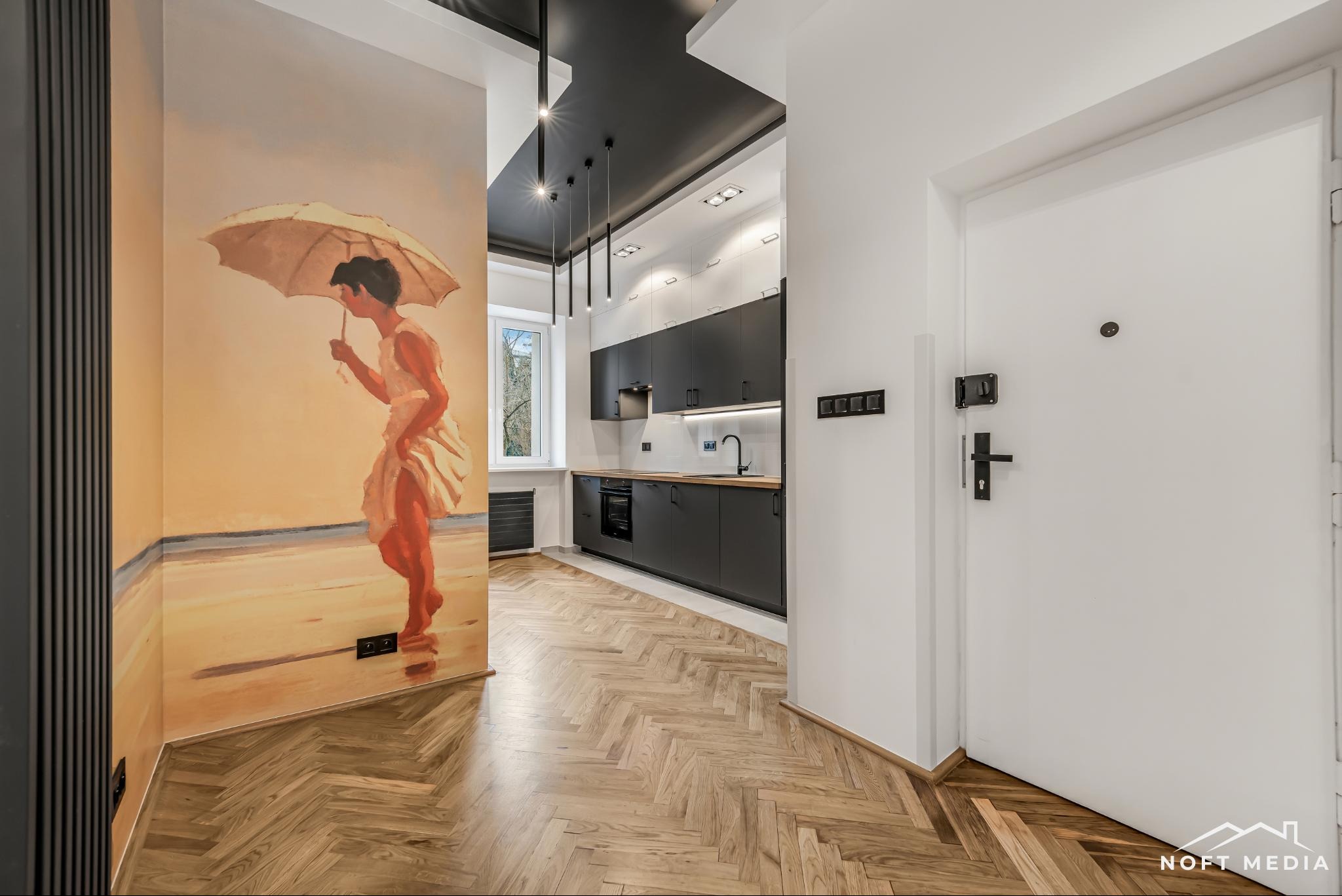 Nowoczesne wnętrze mieszkania z parkietem w jodełkę, czarną kuchnią i malowidłem ściennym przedstawiającym kobietę z parasolem na plaży.