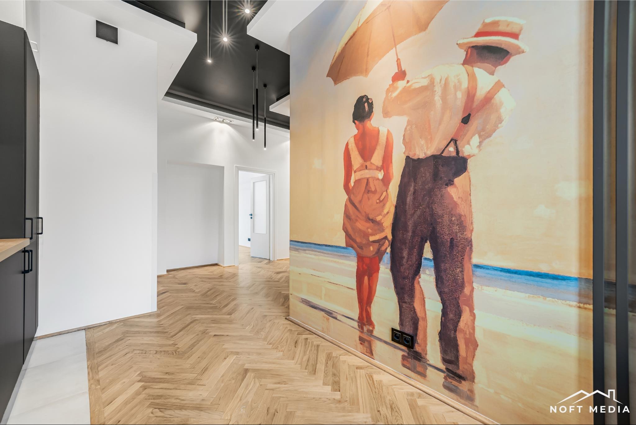 Jasne wnętrze z parkietem w jodełkę i fototapetą z motywem plażowym, przedstawiającą parę osób, z czarnymi akcentami sufitu i oświetlenia.