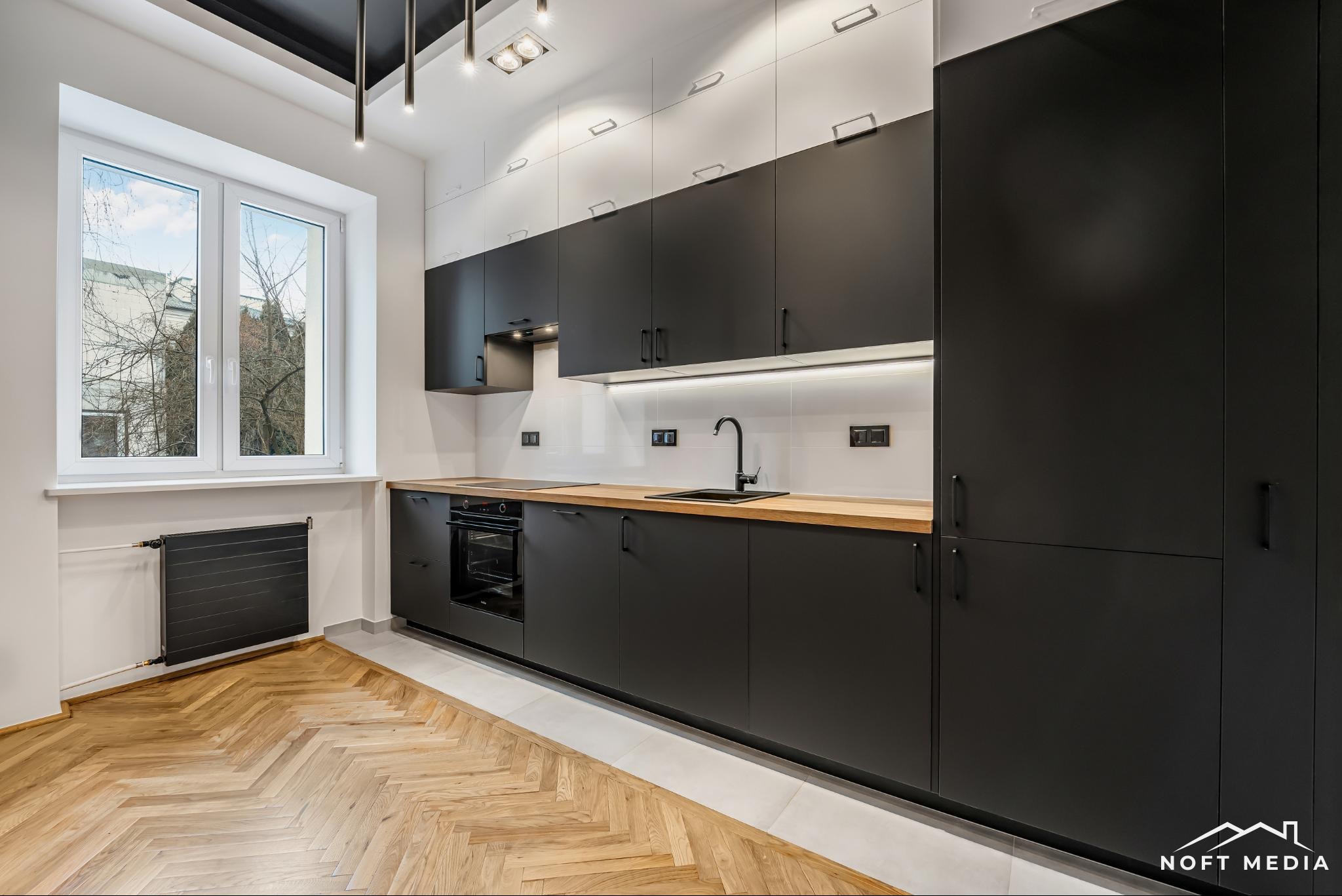 Nowoczesna kuchnia z czarnymi matowymi szafkami, drewnianym blatem, parkietem w jodełkę i grzejnikiem pod oknem.