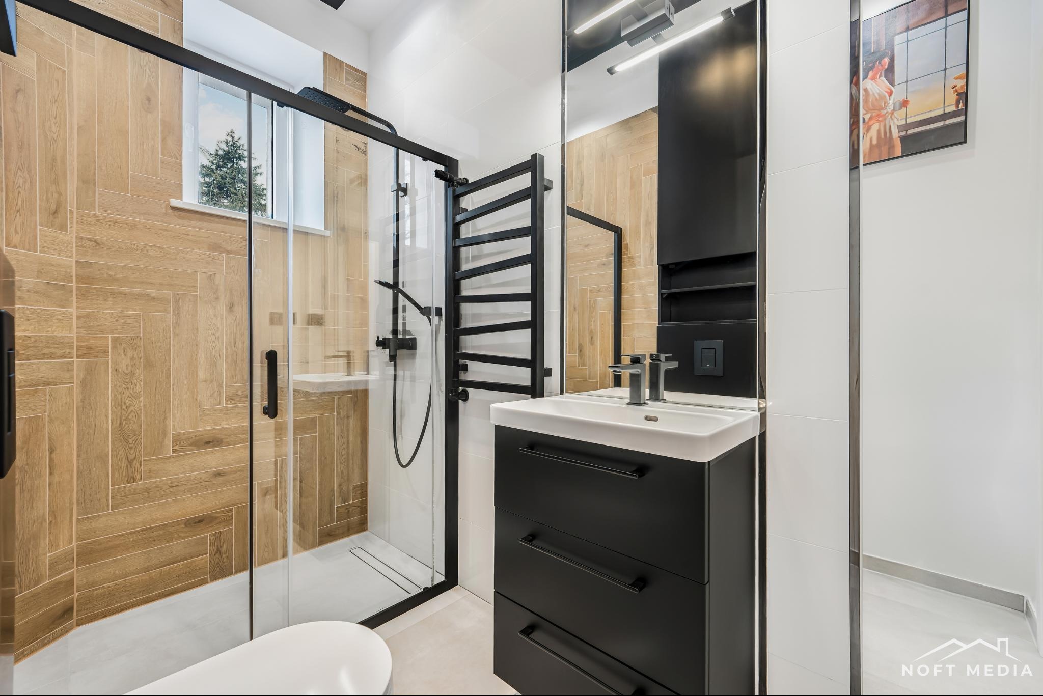 Nowoczesna łazienka z kabiną prysznicową typu walk-in, drewnopodobnymi płytkami ułożonymi w jodełkę, czarną armaturą i szafką umywalkową z trzema szufladami oraz obrazem kobiety w lustrze.