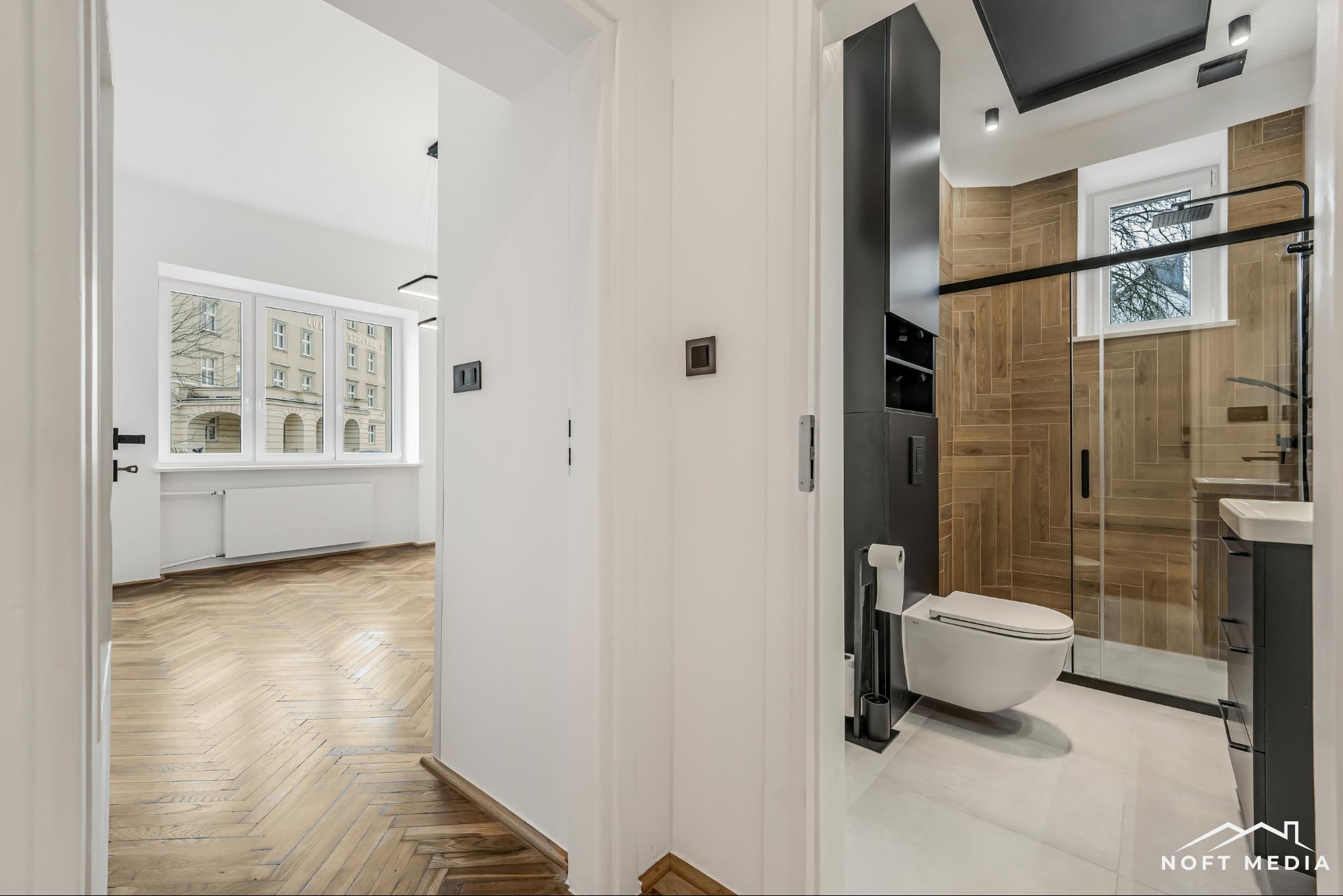 Widok z korytarza na łazienkę z kabiną prysznicową typu walk-in, wiszącą toaletą i umywalką na szafce; w tle parkiet w jodełkę i okno z widokiem na budynek.