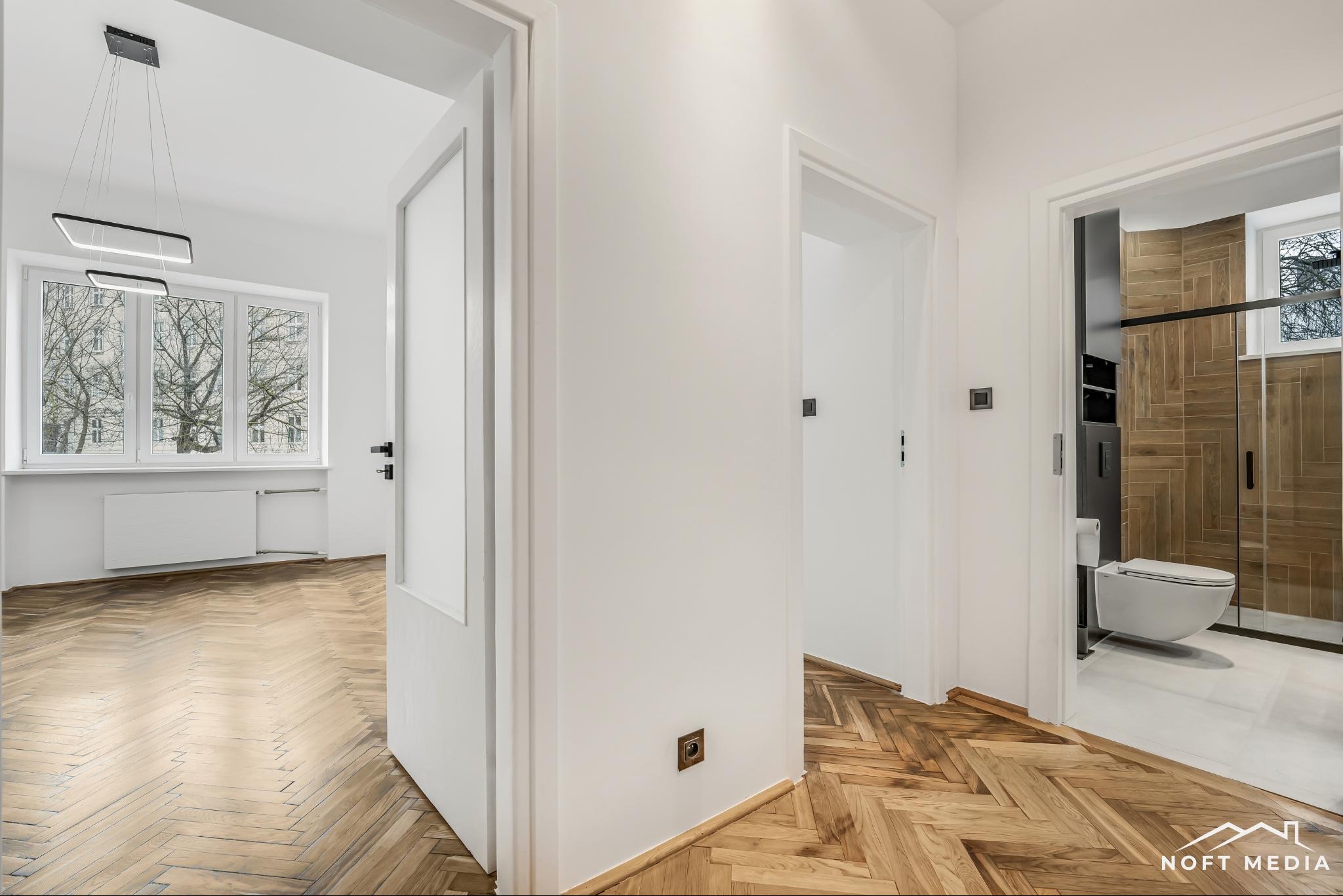 Jasne wnętrze z parkietem w jodełkę, widok na łazienkę z kabiną prysznicową i toaletą, nowoczesna lampa wisząca i duże okno z widokiem na drzewa.